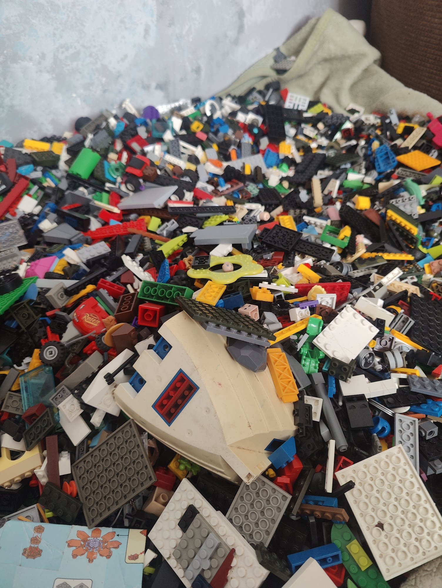 This media file captures a close-up, overhead view of a large, overflowing bin filled with a chaotic jumble of colorful LEGO bricks and assorted toy parts. The scene is dominated by the sheer volume of plastic pieces, with a variety of shapes, sizes, and colors, including red, blue, green, yellow, black, white, and gray. There are also some larger, assembled components, such as a white and beige boat-like structure with blue and red accents, and a gray flat piece with a grid of circular indentations. One of the crops reveals a portion of a LEGO instruction booklet featuring orange and brown LEGO pieces.
The setting appears to be indoors, with a textured, light-colored wall or backdrop visible in the upper left portion of the image. A draped piece of light green fabric is also visible in the upper right, suggesting the bin might be near a piece of furniture or a window covered by a curtain. The lighting is consistent with indoor lighting, with no specific time of day or weather conditions discernible. The overall impression is one of a child's play area or storage space for toys. There are no people visible in the frame. The activity taking place is implied to be the storage or perhaps the aftermath of play with these toys. The location context provided is Yarmolyntsi, Ukraine.