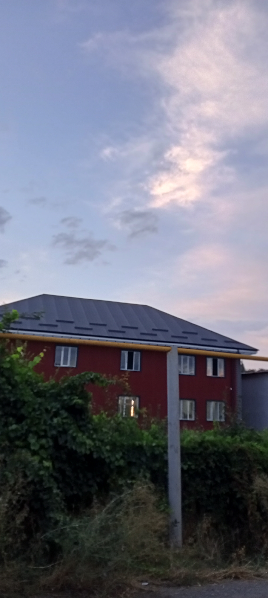 In Sokuluk, Kyrgyzstan, a red building with a dark gray metal roof stands partially obscured by dense green foliage in the foreground. The building has multiple windows, some reflecting the sky. A yellow pipe runs horizontally across the facade, just below the roofline. A concrete pole is positioned in front of the building. The sky above is a pale blue with scattered clouds, some tinged with pink, suggesting either dawn or dusk. The lower portion of the image shows dry grass and some debris on the ground, indicating an outdoor, possibly semi-rural or overgrown urban setting.