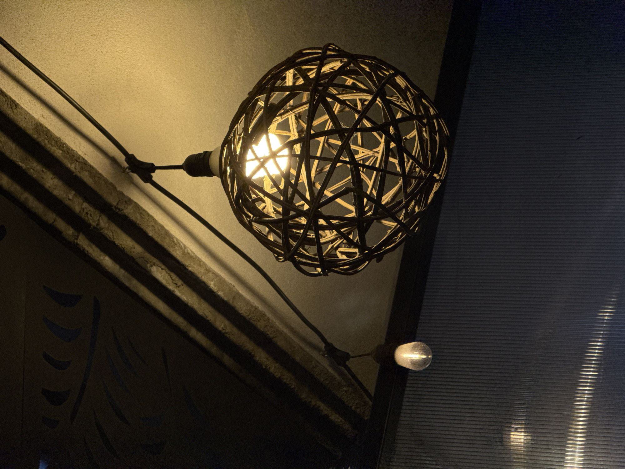 An upward-angled shot captures an illuminated, spherical light fixture, woven from dark, intertwining strips of material, likely rattan or wicker. A bright, warm-toned light bulb glows intensely from within its cage, casting a star-like flare. This main fixture is suspended by a black cable, which extends from the upper left, traverses the image diagonally downwards, and supports a second, smaller, bare light bulb to the lower right.
The primary light illuminates a textured, light-colored wall or ceiling in the upper left portion of the frame, highlighting its rough surface and casting subtle shadows from the cable. Below this, in the darker lower-left quadrant, are indistinct, dark architectural or decorative elements, possibly part of a wall or railing, which also cast prominent shadows from the ambient light.
On the right side of the image, a dark, vertical surface with horizontal ridges is visible, resembling a corrugated panel, window blinds, or a translucent partition. Reflections and faint light patterns are visible on this surface, particularly in the lower right, suggesting another light source or reflections.
The scene appears to be set at night or in a dimly lit indoor space, emphasizing the artificial illumination. The warm glow from the bulbs creates a cozy and intimate atmosphere. No people or visible text are present in the image. The location context is San Andrés Cholula, Mexico.