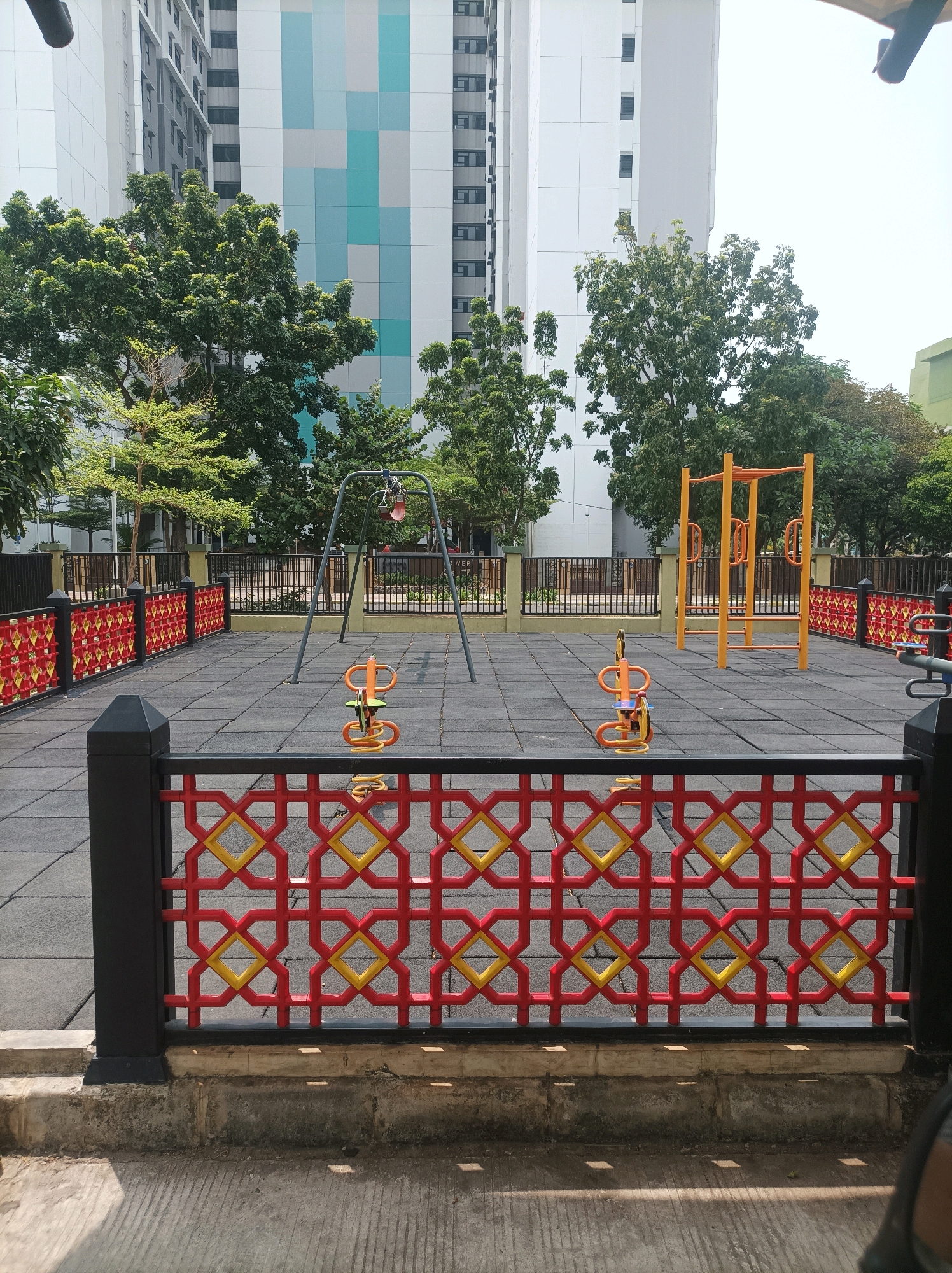 The image depicts an outdoor playground area in West Jakarta, Indonesia. The playground features a swing set, two spring riders, and an orange metal climbing structure. The ground is covered with black rubber matting, and the area is enclosed by a fence with a decorative red and yellow geometric pattern. In the background, there are tall apartment buildings and green trees. The sky is clear and bright, suggesting a sunny day. No people are visible in the image.