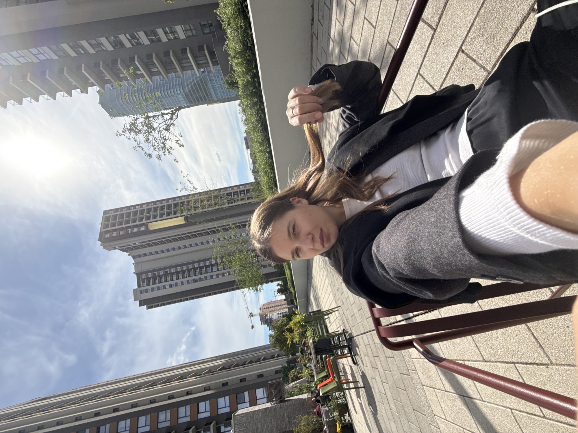 A young woman sits on a metal chair in an urban setting. She wears a white top, a black jacket, and gray sweatpants. Her long brown hair is held in her hand. In the background are tall, modern buildings under a partly cloudy sky. The location is Belgrade, Serbia. The ground is paved with light gray bricks. In the distance, additional chairs and tables suggest a public space or outdoor cafe. A construction crane can be seen among the buildings.