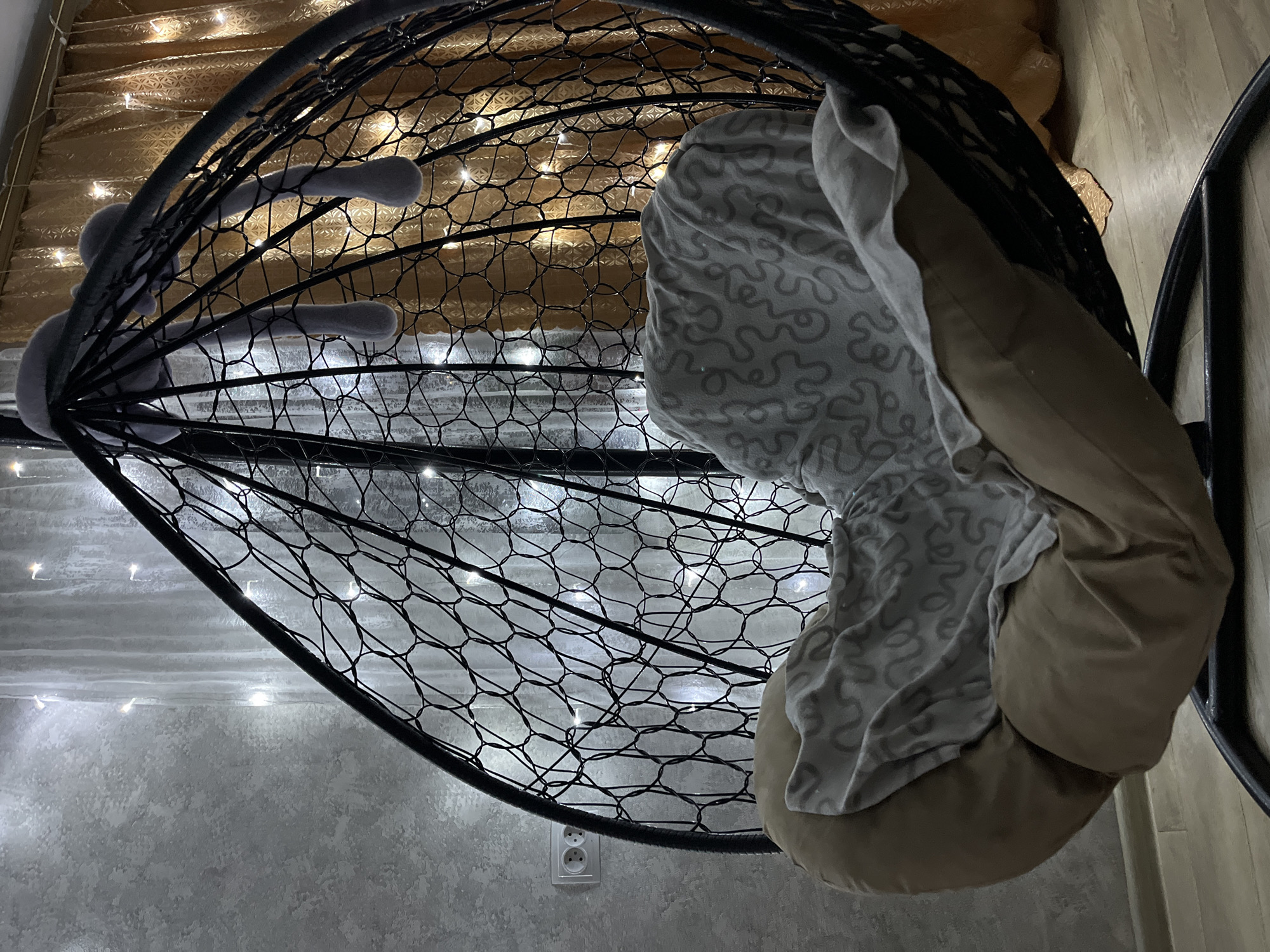 The image shows the interior of a room, possibly in a residential setting. A black, teardrop-shaped hanging chair occupies the central space. The chair is made of a net-like material and is adorned with small, white fairy lights. A plush toy, seemingly a rabbit, is positioned within the chair's netting. Inside the chair sits a tan cushion covered with a patterned gray and white blanket or throw. The background consists of a wall with what appears to be a light-colored texture, and an electrical outlet is visible near the bottom of the frame. The upper portion of the background transitions into a golden-toned surface that might be a curtain or wall covering.
The image does not explicitly provide details that would confirm its location in Novopokrovskaya, Russia. There is no information provided regarding people, actions, or interactions.
