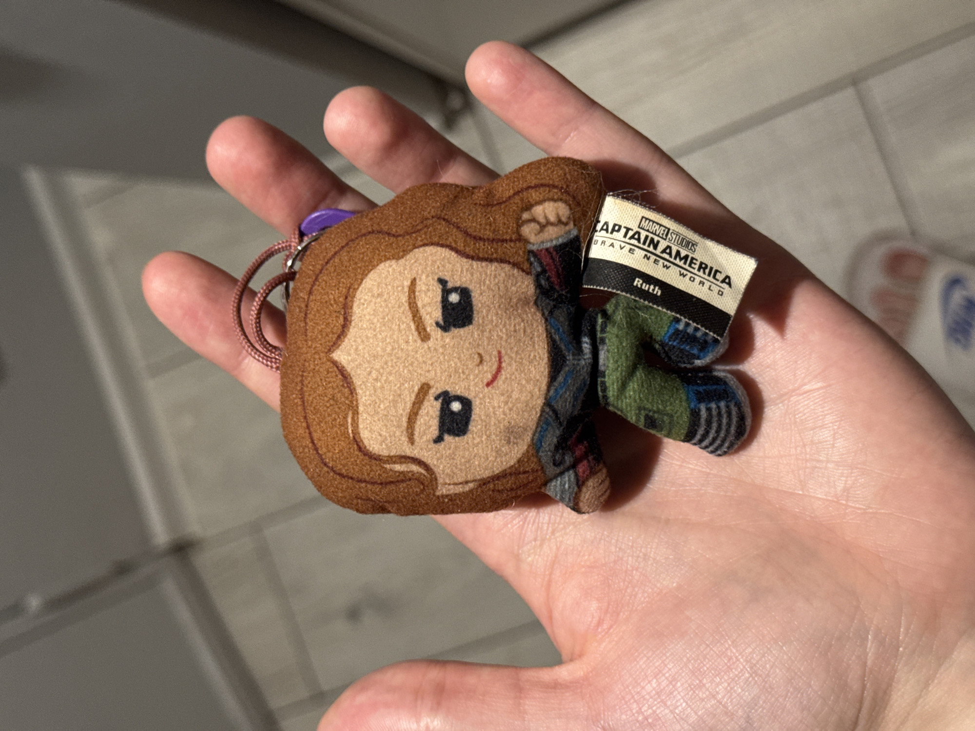 A person's hand holds a small plush keychain of Captain America from Marvel Studios. The keychain is a chibi-style representation of the character, with a round head, large eyes, and simplified facial features. Captain America is depicted wearing his signature costume, including a blue and red outfit with a star emblem, green pants, and striped boots. A small tag attached to the keychain reads "MARVEL STUDIOS CAPTAIN AMERICA BRAVE NEW WORLD Ruth". The background is blurred and appears to be an indoor setting, possibly a home. The lighting suggests a close-up shot taken with a flash.