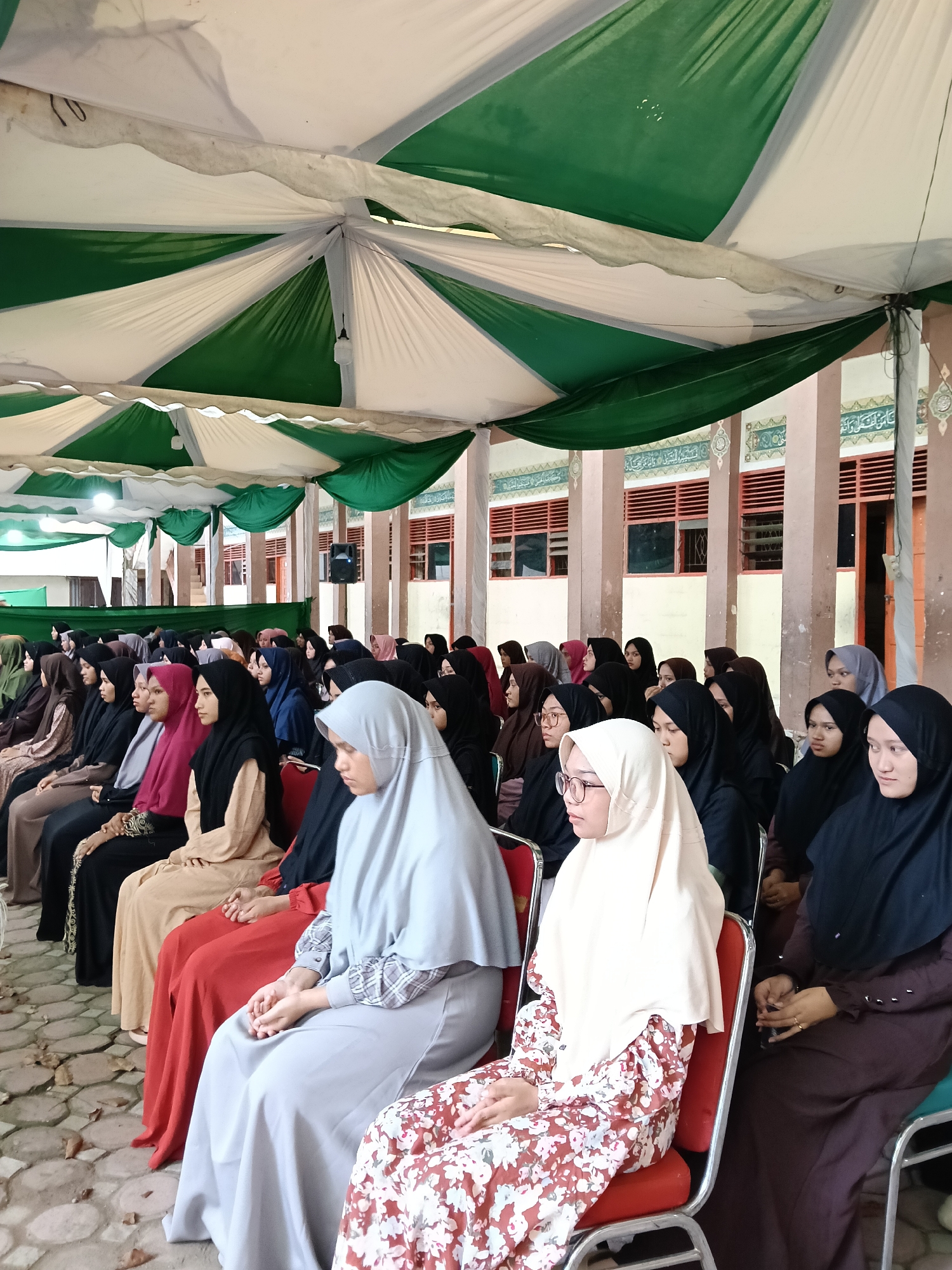 The image depicts a gathering of women seated in rows under a green and white striped tent structure. They are primarily wearing headscarves (hijabs) in varying colors including black, grey, beige, and maroon. The women are dressed in long gowns or dresses. The background shows a building with pillars, windows, and Arabic script above the windows. The ground is paved with a pattern of interconnected circles or cobblestones. Based on the details, it is likely the image was taken in Lhokseumawe, Indonesia.