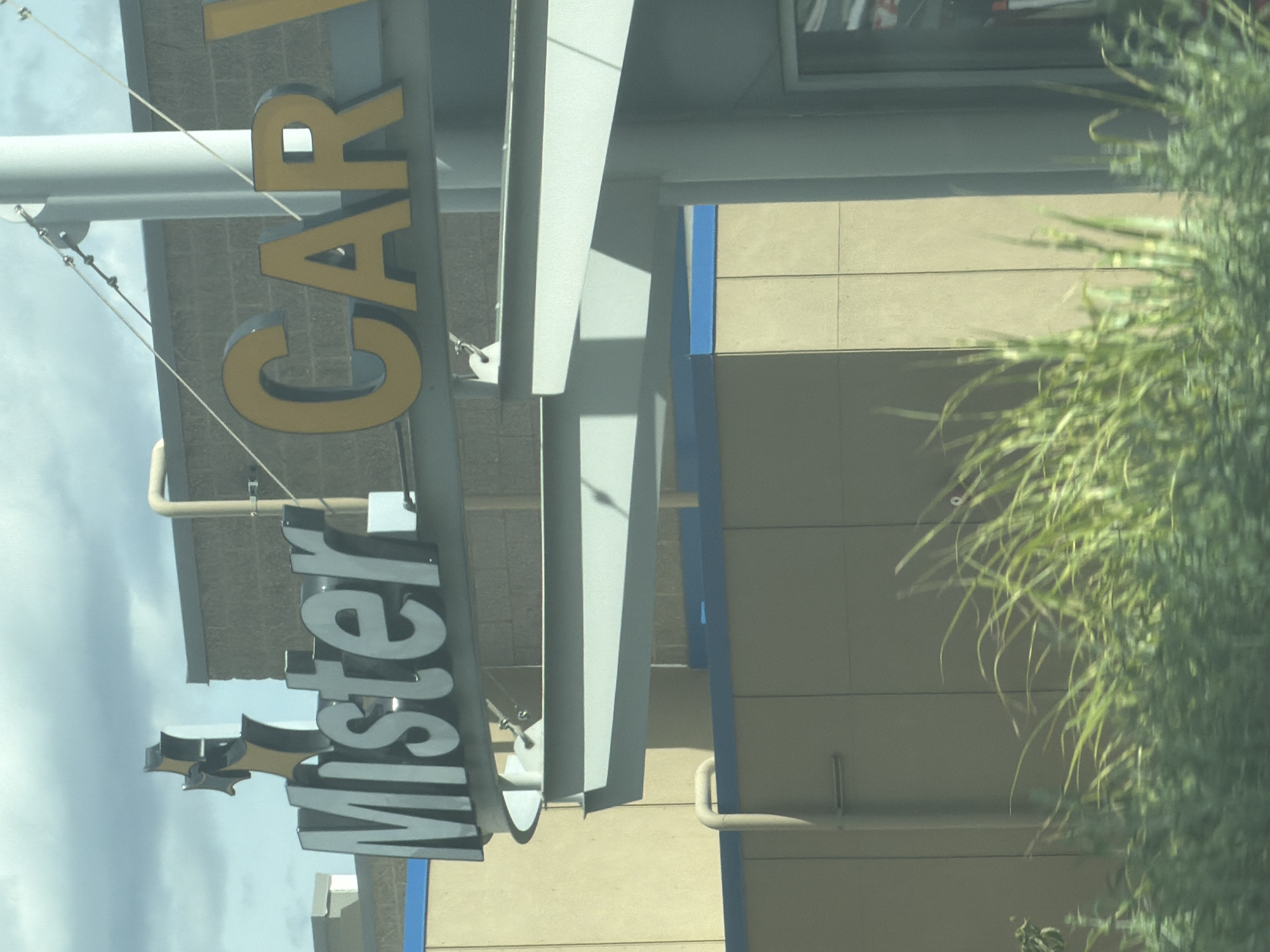 In Pasco, Washington, a "Mister Car Wash" sign is visible above a beige-colored building. The sign features large, grey, block letters spelling "Mister" on the left, and yellow, block letters spelling "CAR" on the right. A stylized, black and yellow graphic resembling a star or a propeller is positioned above the "Mister" portion of the sign. A light-colored pipe runs vertically up the side of the building behind the "Mister" sign. The building is partially obscured by a metal structure and a foreground of blurred green foliage. The sky above is cloudy.