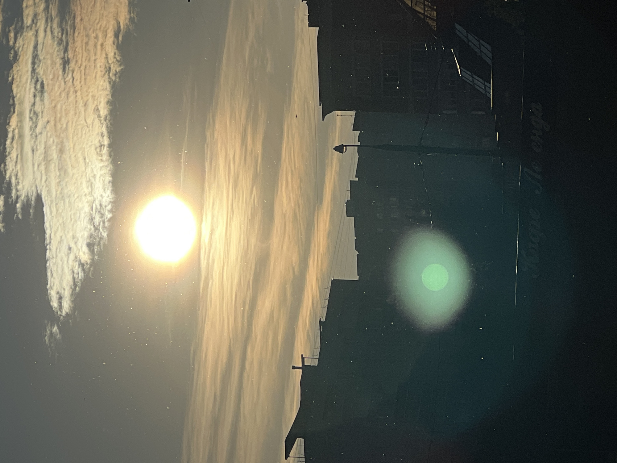 An outdoor photograph captures a cityscape at either sunrise or sunset, viewed through what appears to be a dusty or reflective surface, possibly a window. The image is bathed in warm, golden light. A bright, intense sun dominates the upper-left quadrant, almost glowing white and creating significant lens flare. Long, streaky clouds, illuminated by the sun's low angle, stretch horizontally across the sky, displaying soft golden and orange hues against a muted gray background.
Along the bottom of the frame, the silhouettes of multi-story buildings and a lone streetlight form a dark, jagged horizon line, obscuring any fine details of the structures. A prominent green circular lens flare is visible in the lower-right portion of the image, contrasting with the dark silhouette. Numerous small, bright specks, likely dust or reflections on the lens or viewing surface, are scattered throughout the frame.
Notably, reversed text, possibly Cyrillic, is faintly visible on a dark surface in the bottom-right corner. The text appears to read "треба до сюда", which translates to "need to here" or "must go here" in Ukrainian. This detail, combined with the general appearance, suggests the photo was taken as a reflection on glass. Given the location context of Khimki, Russia, the Ukrainian text is a curious element, perhaps originating from an object inside the reflected environment.