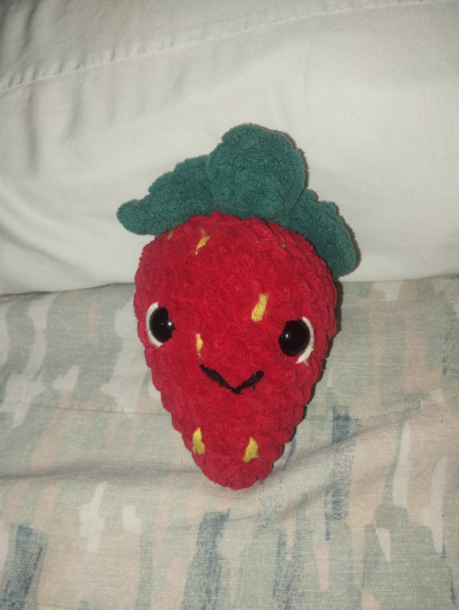 A plush toy strawberry with green leaves, a red body with yellow seed markings, and a stitched black smile and plastic eyes is resting on a patterned fabric surface, with a solid white fabric in the background. The location is not discernible from the image.