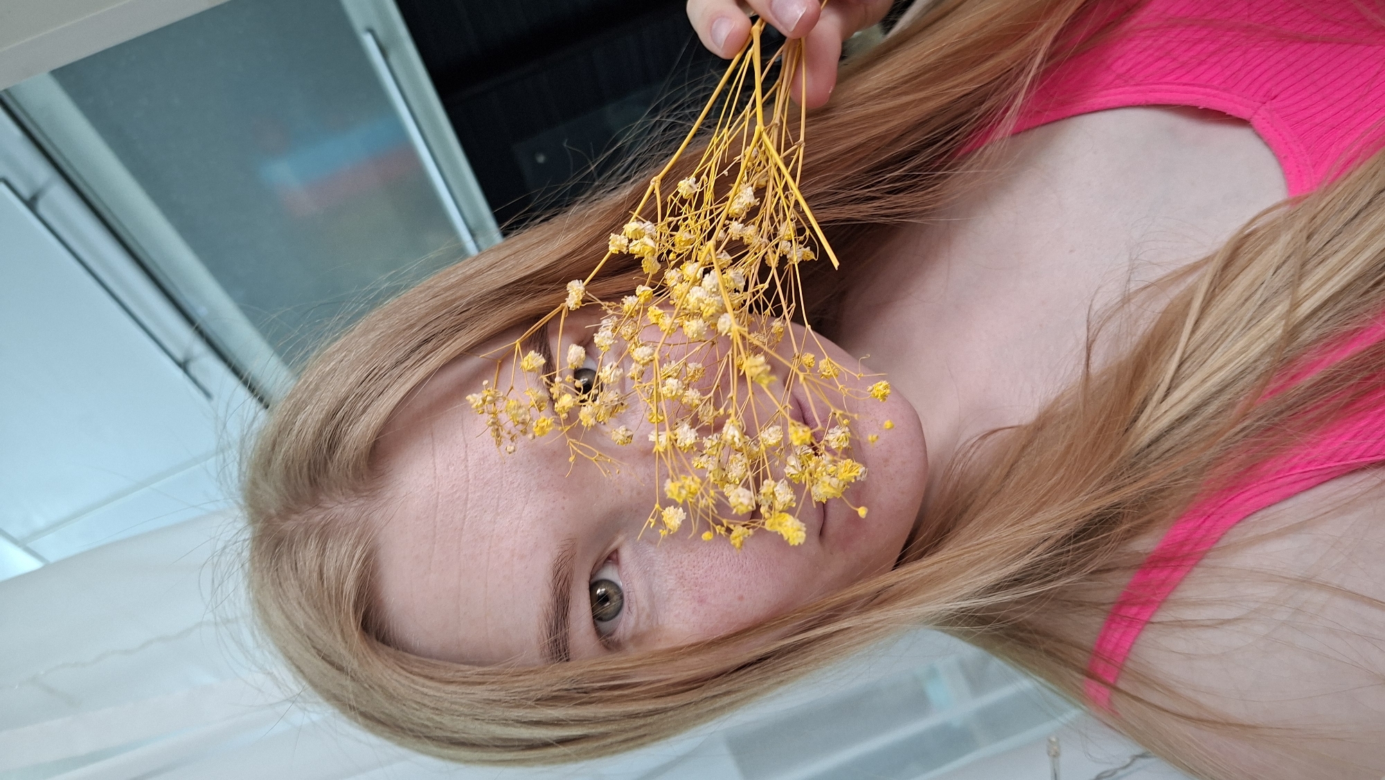 A blonde woman in a pink top is holding a bunch of small yellow flowers in front of her face. She has green eyes and fair skin. The background is blurred, but appears to be an indoor setting. The image may have been taken in Chaikovsky, Russia.