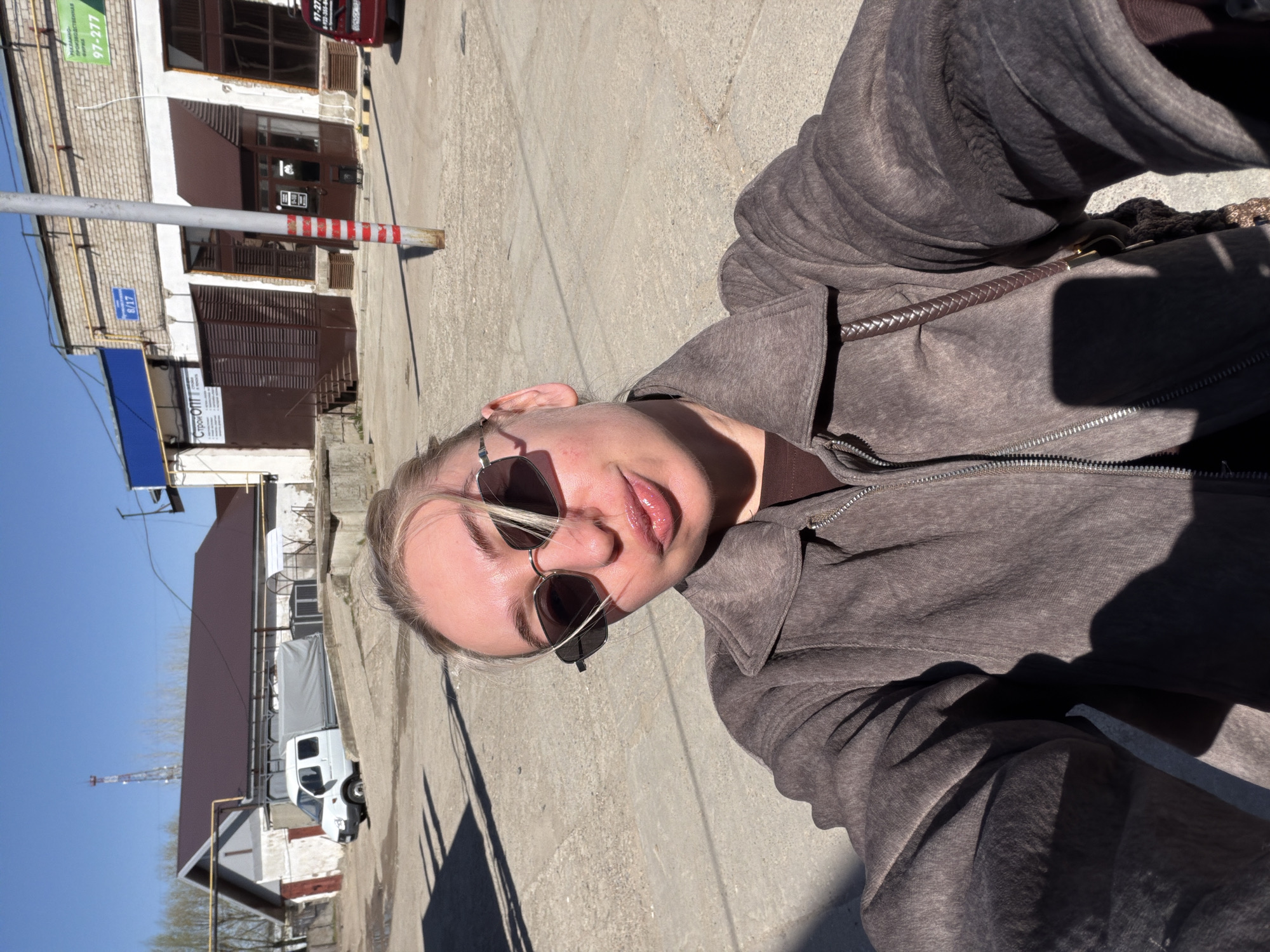 A woman wearing sunglasses and a brown jacket is taking a selfie in Chaikovsky, Russia. The photo shows her face and upper body. In the background is a street with buildings, a white van, and a red and white striped pole. The sky is blue.