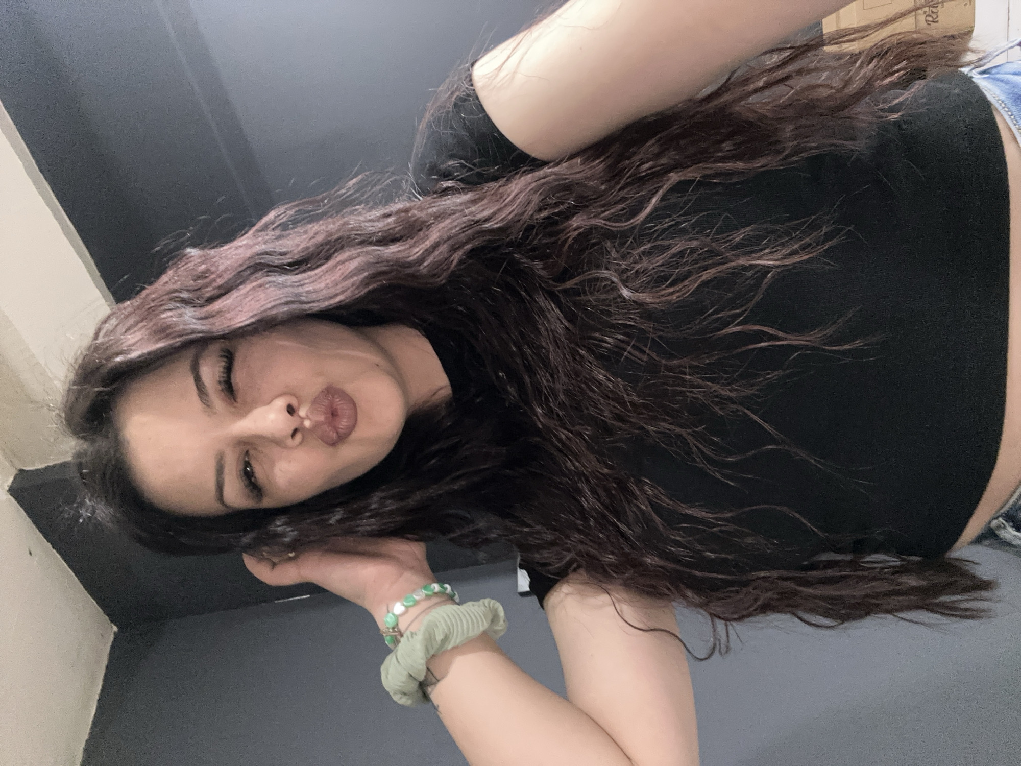 A selfie of a young woman with long, wavy brown hair, wearing a black t-shirt and blue jeans, is shown. She is pouting her lips and looking into the camera. The woman is wearing a green scrunchie and a beaded bracelet on her left wrist. The background consists of gray walls and a box in the upper right corner. The image does not provide information about the city of Sliven, Bulgaria.