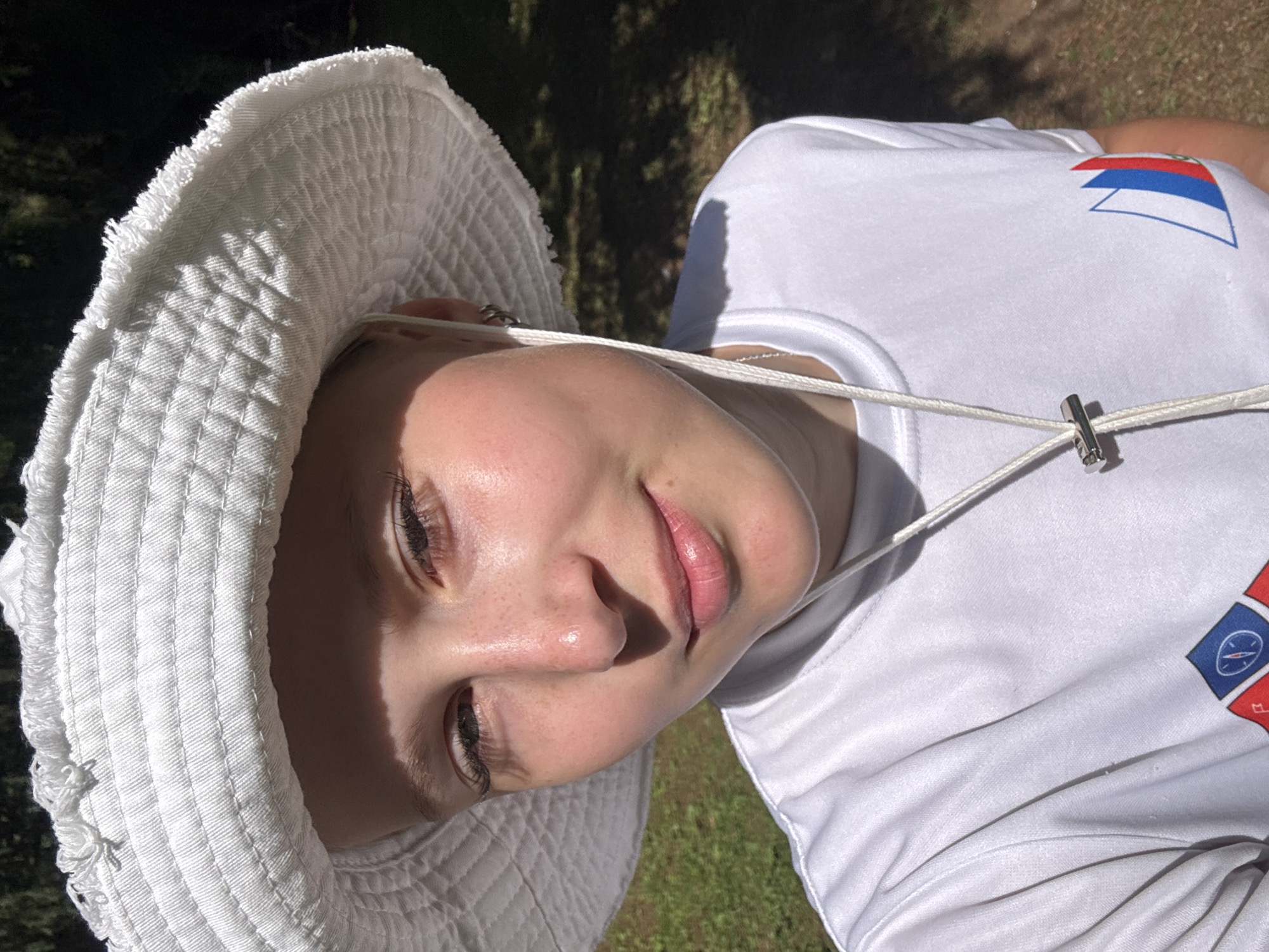 The image is a close-up, low-angle portrait of a young person wearing a white bucket hat and a white t-shirt with a logo on the right shoulder. The person has light skin with freckles and is looking towards the camera with a slight smile. Their eyes are open, and the light is catching their face, creating some shadows. A white drawstring from the hat is visible across their chest, secured by a metal clasp. The t-shirt has a design with red, white, and blue elements, possibly a flag or emblem, on the right shoulder. Another emblem, featuring a compass-like design within a red and blue square, is visible on the chest.
The setting appears to be outdoors, with green grass and dark foliage in the background, suggesting a park or natural area. The lighting indicates it is daytime, likely a sunny day given the strong shadows. The overall mood is relaxed and informal. The user-provided location context is Сергино, Russia. There is no visible text in the image, other than the emblem on the t-shirt which is not legible as text.