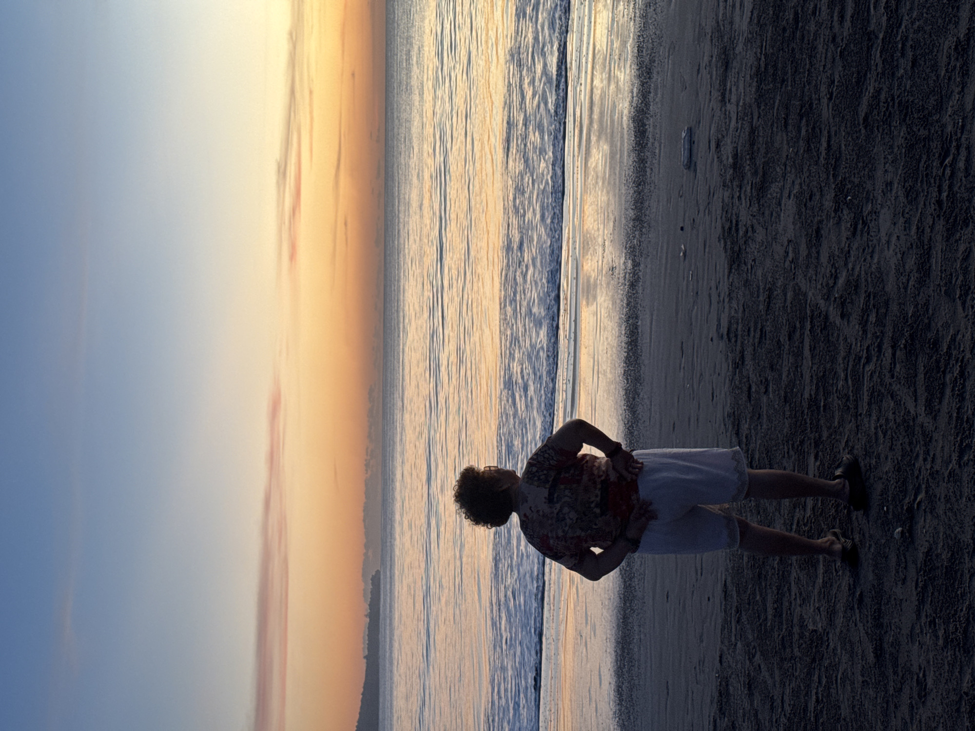 The image shows a person standing on a beach at sunset, possibly in Kuta Selatan, Indonesia. The person is silhouetted against the colorful sky and ocean. The sand is dark and appears wet. The person is wearing a patterned shirt and light-colored shorts. The sky is a gradient of colors, from blue to orange. The water is reflecting the colors of the sky.