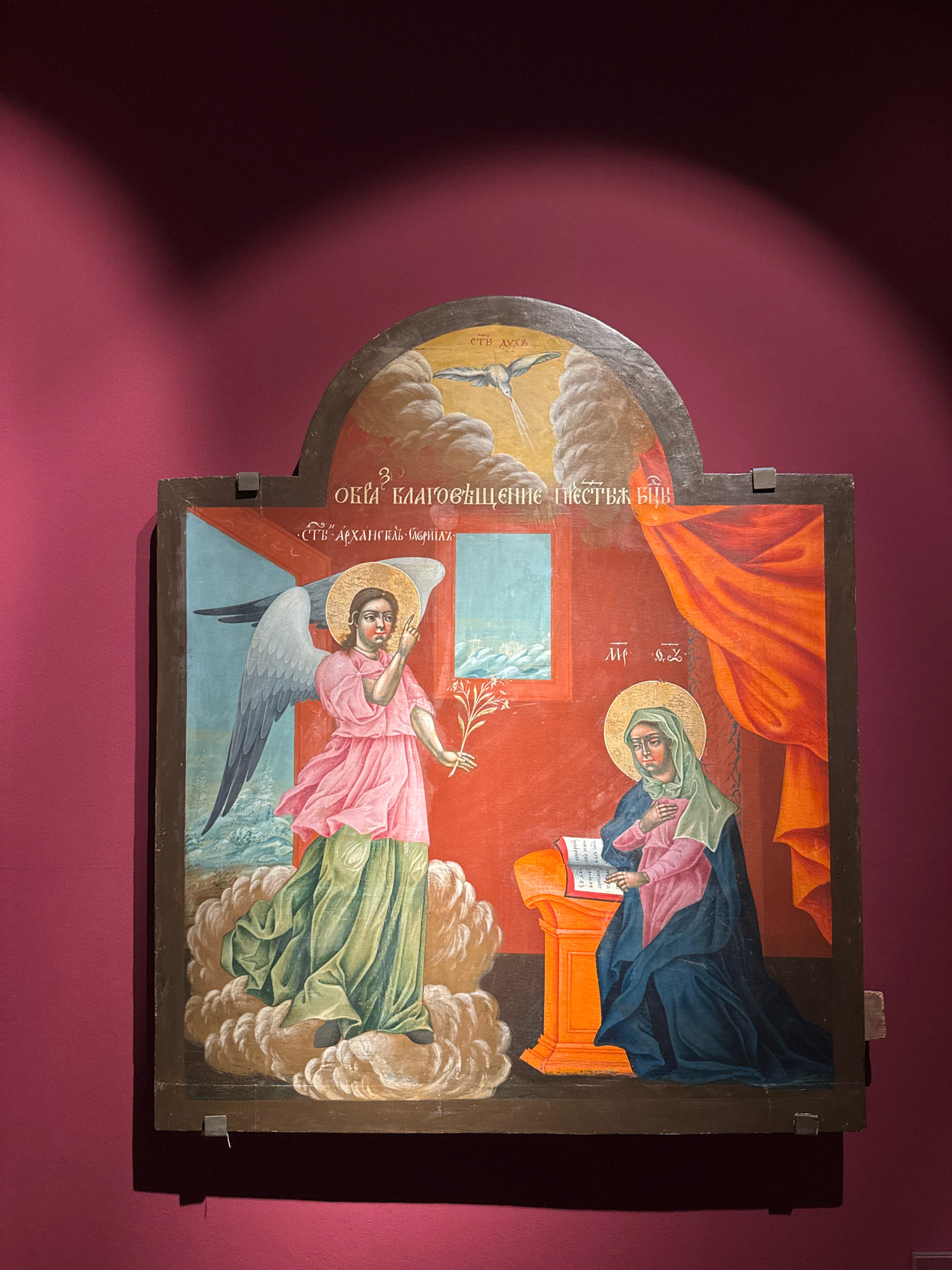 An Eastern Orthodox icon, likely depicting the Annunciation, is displayed against a plain, dark pink wall. The icon is housed within a dark wooden frame, and the top of the frame is arched. The scene within the icon is divided into two main registers and a smaller upper section.
The upper section features a dove, representing the Holy Spirit, descending from a cloud-filled sky. A radiant light emanates from the dove, and some faint lettering is visible above it.
The lower section of the icon depicts two figures. On the left, an angel with large grey wings, wearing a pink tunic and a green robe, stands on a cloud and holds a lily stalk. The angel's halo is gold, and its gaze and raised finger suggest communication. The background behind the angel is a light blue sky with a suggestion of water and distant structures.
On the right, a woman, presumably the Virgin Mary, sits before an orange lectern with a book open on it. She wears a blue cloak over a pink garment and a green headscarf. Her left hand is placed on her chest, and her gaze is downcast. A golden halo surrounds her head. The background behind her is a solid red, with a depiction of orange drapery on the far right. White Cyrillic lettering is visible in several locations on the icon, including above the angel and near the woman.
The icon is secured to the wall with visible metal brackets at the bottom. Shadows from an unseen light source are cast on the wall behind and to the left of the icon, with a distinct gradient of darkness from top to bottom. The overall impression is of a religious artifact presented in a museum or gallery setting.