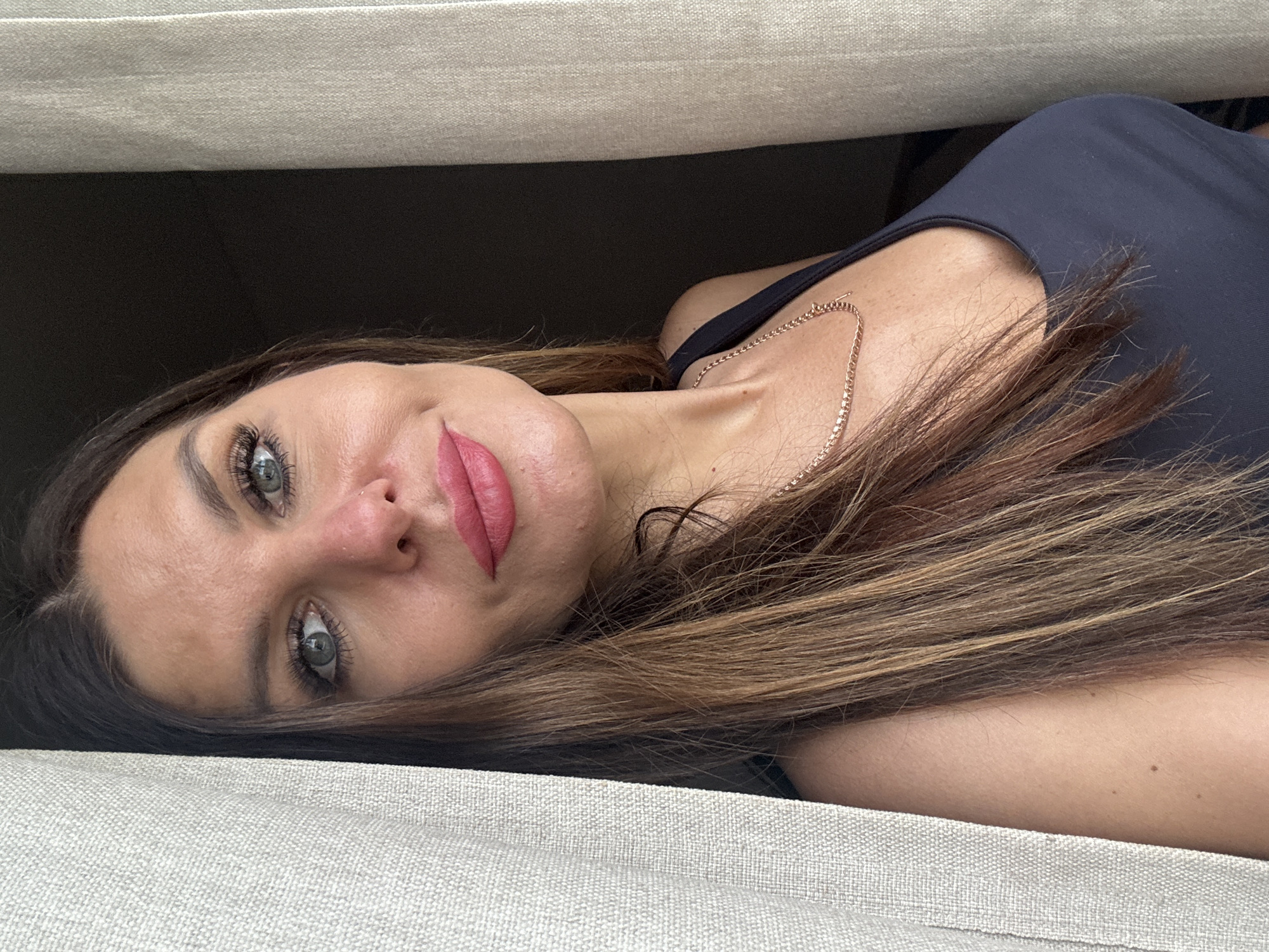 A woman with brown and blonde hair, wearing a black top and a gold chain necklace, lies on a beige textured surface with a dark background. Her head is tilted, revealing pink lipstick and grey eyes. The scene is indoors, with the textured surface possibly being a sofa or bed. The location in Chaikovsky, Russia is unknown.