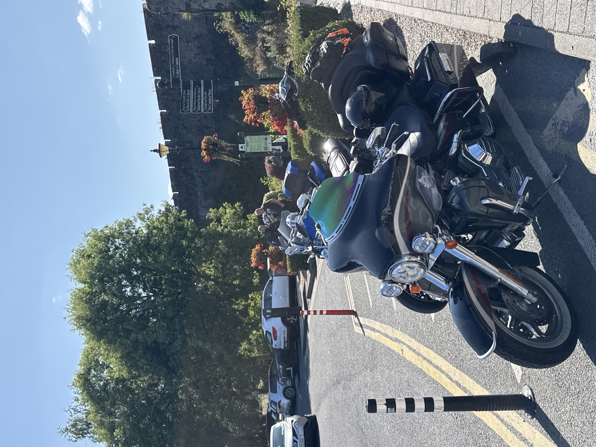 A daylight, eye-level, medium shot captures a parked Harley Davidson motorcycle in the foreground on a paved surface with yellow dividing lines. The motorcycle is black with silver accents and has a tinted windshield with a rainbow sheen. Behind it, other motorcycles and several cars are parked in a lot bordered by a stone wall and lush green trees. A signpost attached to the wall displays directions to "Cahir Castle," "St. Mary's R.C. Church," "Medieval Parish Church," and "The Convent of Mercy," indicating the location is Cahir, Ireland. The sky is clear blue with a few wispy clouds. Several people are visible in the background, some standing near the motorcycles and others walking further back. A lamppost with a decorative planter hangs above the stone wall. The scene appears to be a popular tourist area or a gathering spot.
