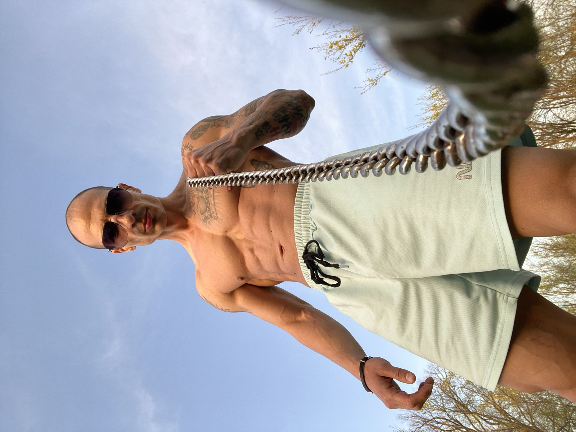 The image displays a fit, shirtless man captured from a low-angle perspective against a clear blue sky. The man has a shaved head and wears dark sunglasses, looking directly at the camera with a serious expression. His body shows visible musculature, including well-defined abs and arms, which are extensively covered in various tattoos.
He wears light mint-green shorts with a black drawstring. A faint, raised logo, which appears to be "IN", is visible on the left leg of his shorts. On his left wrist, he has a dark band or bracelet.
The man is holding a long, flexible, metallic coiled object horizontally across his upper body, with his hands gripping it on either side. This object, resembling a large spring or flexible pipe, is a prominent feature in the foreground.
The background is dominated by a bright blue sky with subtle cloud formations, indicating an outdoor setting during daytime. Bare branches of trees with sparse yellow leaves are visible in the upper corners, suggesting the photograph was taken in either early spring or late autumn. The overall scene conveys a strong, posed image of the man. The location context indicates Pleven, Bulgaria, although no specific landmarks are visible.