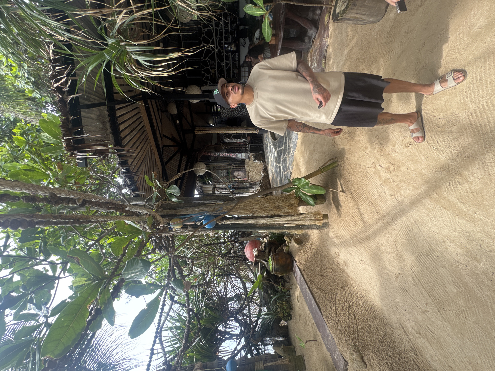 A man stands on a sandy surface near an establishment in ตำบลเกาะพะงัน, Thailand. The man wears a light-colored t-shirt, dark shorts, and sandals. He has visible tattoos on his arms and legs. The establishment behind him features a thatched roof, wooden beams, and decorative elements, including string lights and possibly hanging ornaments. Tropical foliage and trees are visible in the surrounding area.