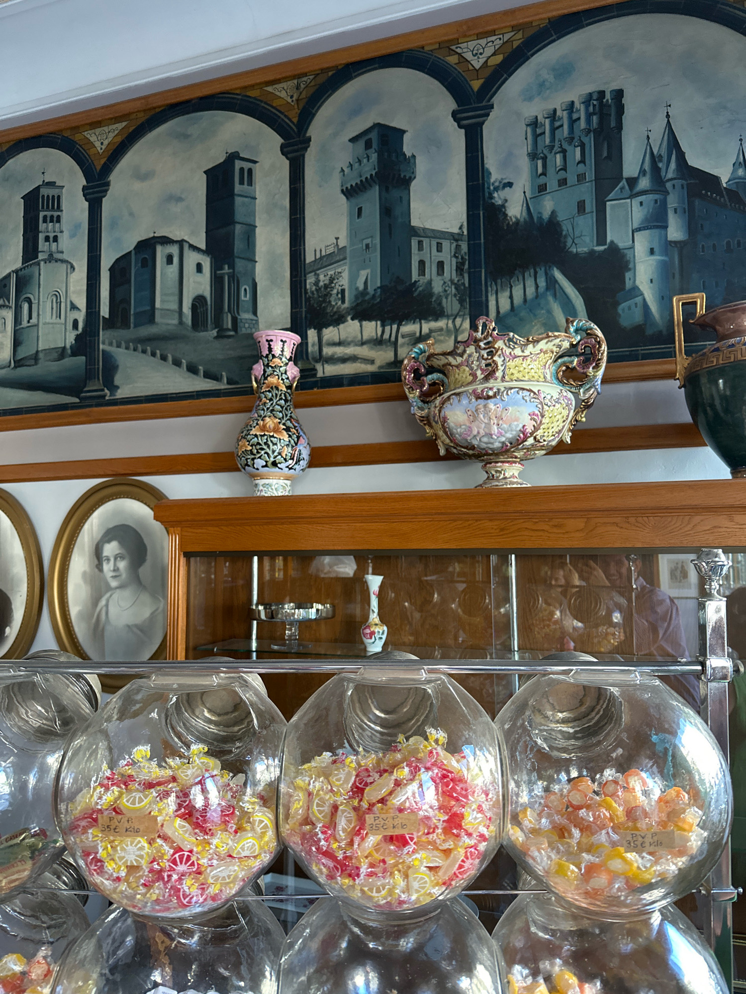 The image depicts the interior of a shop, likely a confectionary or bakery, in La Lastrilla, Spain. A decorative mural of arched cityscapes dominates the upper portion of the scene. Below the mural, a wooden shelf displays several ornate decorative items, including two ceramic vases and a large, intricately decorated bowl. To the left, two framed portraits of women are visible on the wall. The lower section of the image features multiple large, clear glass jars filled with assorted candies. Each jar has a small handwritten label, with one clearly legible reading "P.V.P. 35 € Kilo". The overall impression is one of a charming, possibly antique, establishment.