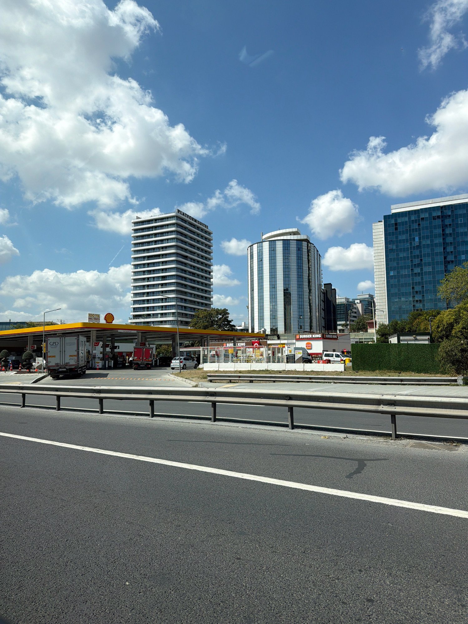 The image shows a highway in Gungoren, Turkey, with several modern buildings and a gas station in the background. The sky is blue with scattered white clouds. On the left, there is a Shell gas station with trucks and cars. Behind the gas station, a tall white building with the text "PLATFORM SUITES" is visible. In the center, a curved glass building and another taller blue glass building are prominent. The foreground shows the asphalt of the highway with a white line marking the lane. A metal guardrail separates the highway from the area behind it.
