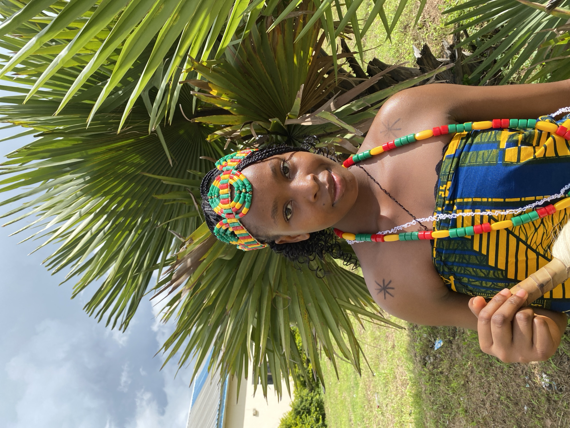 This is a portrait of a Black woman in Wang, Nigeria, outdoors during the daytime. She is wearing a colorful, traditional outfit consisting of a blue and yellow patterned dress and a beaded headband and necklace in red, green, and yellow. The beads are strung in repeating patterns. Her hair is styled in braids, and she has a tattoo of a starburst on her upper arm. She is looking towards the camera with a gentle smile.
The woman is positioned in front of lush green palm fronds, with the sky and some clouds visible in the background. A building with blue accents and some greenery can also be seen in the distance. The scene suggests a warm, possibly humid, day. The woman appears relaxed and happy. There is no visible text in the image.