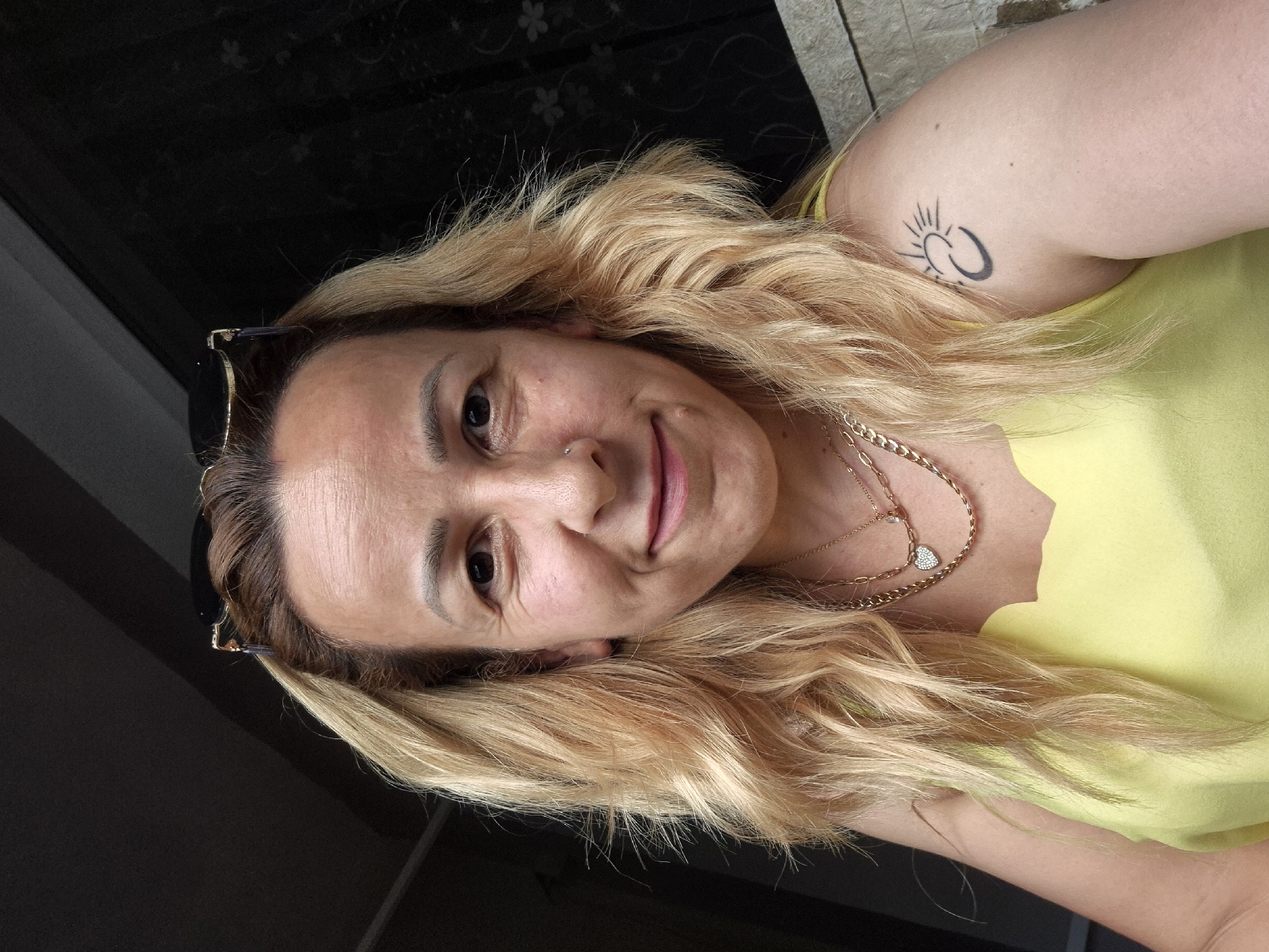 A close-up, head and shoulders shot shows a woman with blonde, wavy hair and sunglasses perched on her head. She is wearing a yellow top and multiple gold necklaces, one of which has a heart pendant. A crescent moon and sun tattoo is visible on her left shoulder. The background is dark and appears to be an interior setting with some dappled light. The woman is smiling. The image was taken in Unknown, Unknown.
