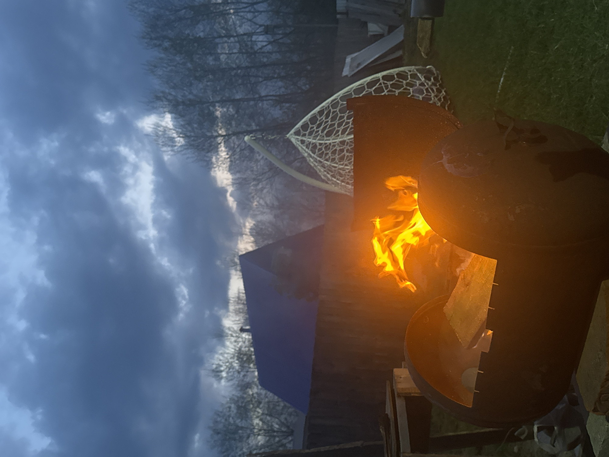 A wood-burning stove is ablaze outdoors. The sky is overcast. A net is in the background. The location cannot be determined as being in Chaikovsky, Russia, based on the visual information.