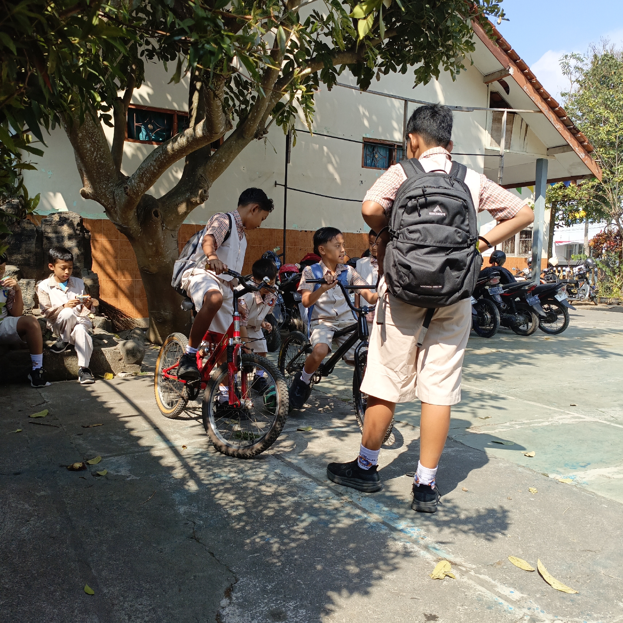 In Salatiga, Indonesia, on a sunny day, four school-aged boys, all wearing matching uniforms consisting of light-colored shirts, vests, and shorts, are gathered outdoors. One boy, standing and facing away from the camera, wears a black backpack. Two other boys are on bicycles, one red and one black, with one of them appearing to be talking. The fourth boy is seated on a low stone wall, looking down at something in his hands. In the background, a white building with a brown-tiled roof is visible, along with several parked motorcycles. The ground is paved, and a large tree with dappled sunlight filtering through its leaves casts shadows across the scene. Fallen leaves are scattered on the ground.