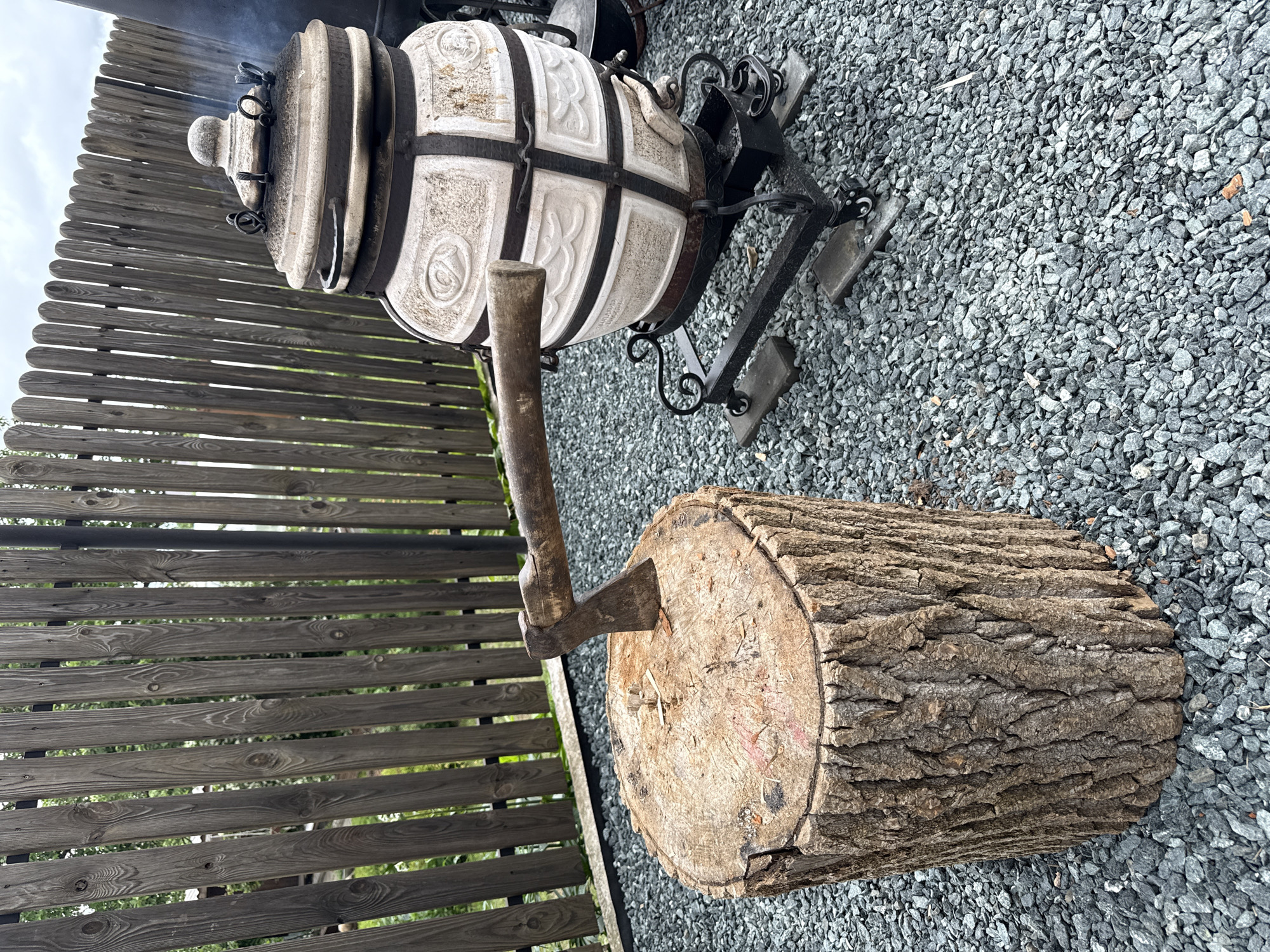 This image, taken in the city of Chaikovsky, Russia, depicts an outdoor utility area. The ground is uniformly covered with small, irregular gray gravel. In the left foreground, a substantial wooden log, approximately 60-70 cm tall and 40 cm in diameter, stands vertically, serving as a chopping block. Its sides retain rough bark, while the top surface, showing concentric growth rings and light splintering, has an axe embedded in it. The axe's dark, weathered wooden handle extends horizontally from the log, with its metal head firmly wedged into the wood.
To the right, a large, light-colored ceramic tandoor oven, approximately 1 meter tall, rests on a black, ornate wrought-iron stand equipped with small wheels and what appear to be supporting blocks beneath them. The tandoor's exterior features decorative relief patterns and is reinforced with dark metal bands. A small amount of white smoke is visible rising from the tandoor's top opening, indicating recent or ongoing use. A small metal tray for ash collection is visible at the base of the stand.
In the background, a weathered wooden fence, composed of vertical planks, forms a boundary. Beyond the fence, green foliage from trees and shrubs is visible under an overcast, light gray sky. No individuals are present in the scene.