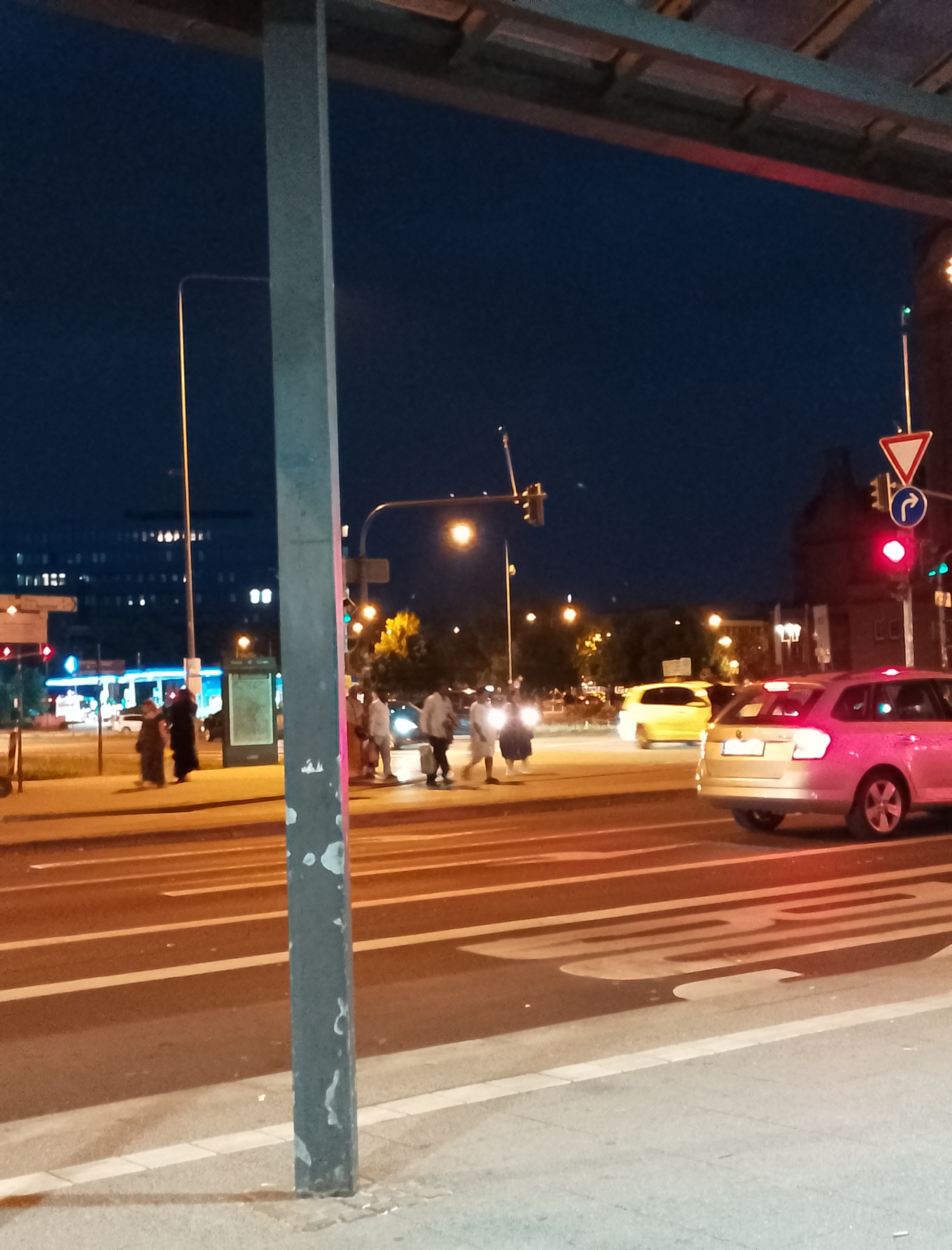 The image depicts a nighttime street scene in an urban environment. A concrete pillar, partially obscured by the foreground, stands to the left. Beyond it, a multi-lane road is visible, with painted lane dividers. Several cars are present, including a silver station wagon in the right foreground and a yellow hatchback further back. Pedestrians are seen crossing the street at a designated crosswalk, some appearing to be walking in groups. Traffic lights are visible, with a red light illuminated. In the background, buildings with illuminated windows suggest a commercial or residential area. The sky is dark blue, indicative of nighttime. The overall impression is that of a busy intersection at night.
