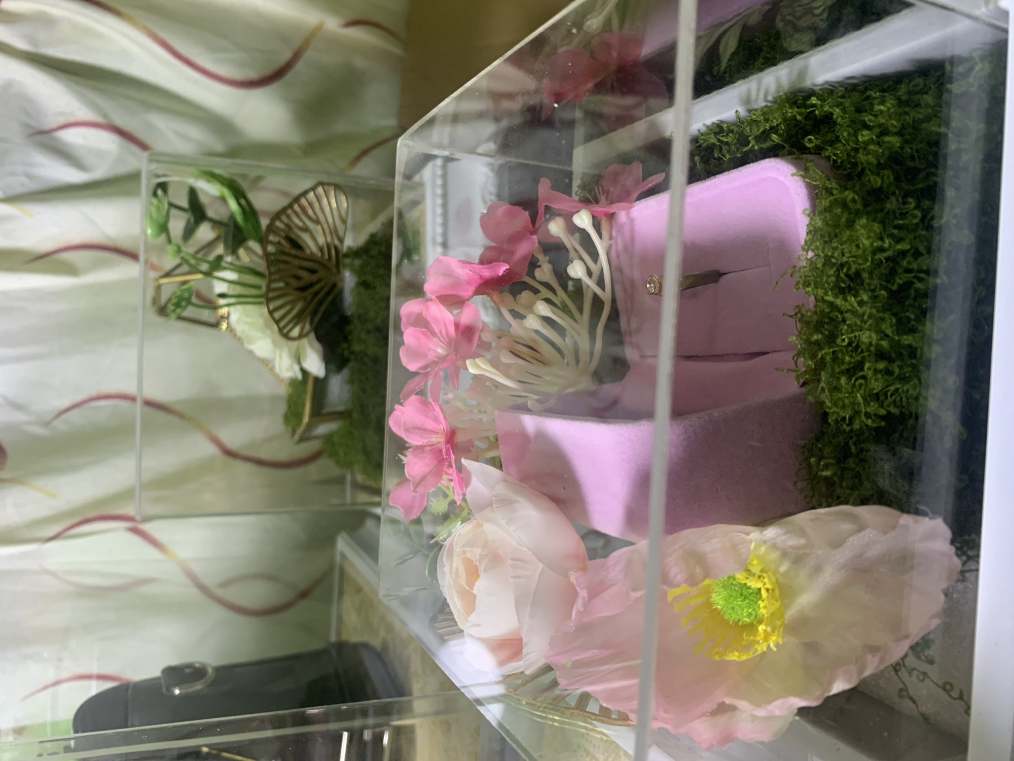 The image shows a close-up view of a clear acrylic box containing decorative items. Inside the box, there are several artificial flowers in shades of pink and white, arranged on a bed of green moss. A pink velvet ring box is also visible, with a ring on display. In the background, a sheer curtain with a red-toned wavy pattern is present, and part of a black object, possibly a bag or purse, can be seen on the left. The scene is indoors, and the lighting suggests an ambient setting. The image does not provide any information about the city of Kuala Pembuang, Indonesia, or any specific location within it.