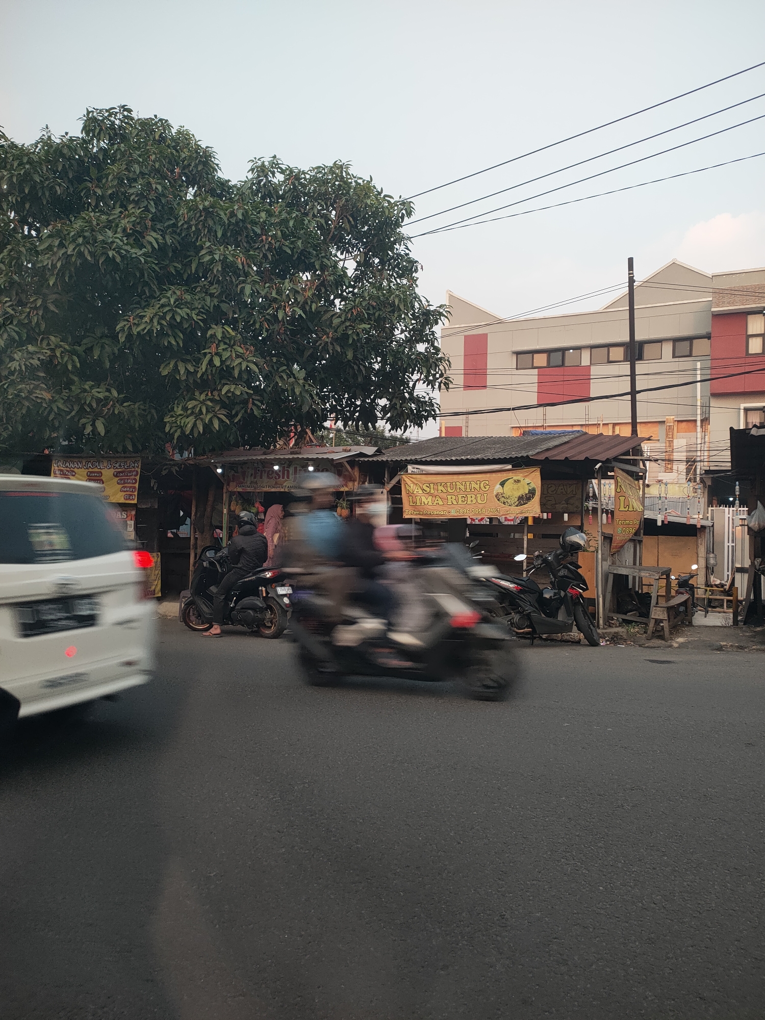 The image depicts a street scene in Cimahi, Indonesia, at what appears to be dusk. The foreground is dominated by a paved road where several motorcycles are in motion, their forms blurred due to movement. A white car is partially visible on the left, also in motion. In the mid-ground, a row of small food stalls lines the street, characterized by corrugated metal roofs and various banners. One banner prominently displays "NASI KUNING LIMA REBU" and a phone number. People are seen near the stalls, some standing and others on motorcycles. A large tree with dense foliage stands to the left of the stalls, providing shade. Behind the stalls, a modern building with red and gray accents is visible, featuring multiple windows. Electrical wires crisscross the sky above the scene. The overall atmosphere is that of a busy street in a commercial area, with the activity of daily commerce and transportation.