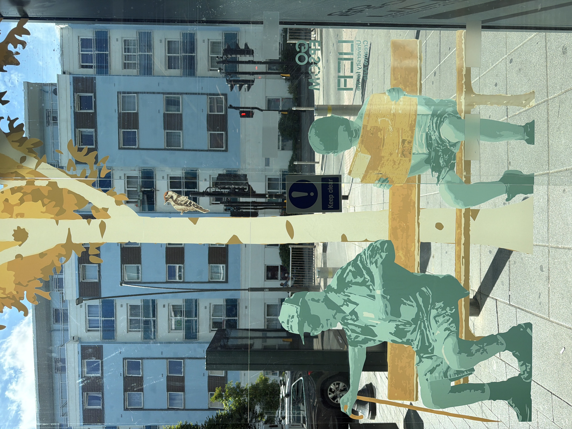 The image shows a street scene viewed through a glass window, likely in Epsom and Ewell, United Kingdom, as suggested by signage visible on the glass. The foreground features a stylized graphic art installation depicting two figures on a bench. On the left is a figure of an older person wearing a cap, sunglasses, and casual clothing, holding a walking stick. On the right is a younger figure, appearing to be a child, reading a book or newspaper. Behind the figures, a tall, stylized tree trunk with yellow leaves is visible, also part of the art.
Through the window, a modern, light blue apartment building with brown accents is prominent. The building has multiple stories, each with numerous windows and balconies with blue railings. A street with traffic lights, lampposts, and parked cars is visible in the middle ground. One traffic light shows a red signal. A sign with an exclamation mark and the text "Keep clear" is also visible on a post. Reflections on the glass obscure some details of the background, including a black car and a fence. Text, partially obscured and reversed, indicating "UNIVERSITY FOR THE CREATIVE ARTS" and "EPSOM" is visible on the glass in the lower right portion of the image. The pavement in front of the bench is paved with grey tiles. The sky above is blue with scattered white clouds.