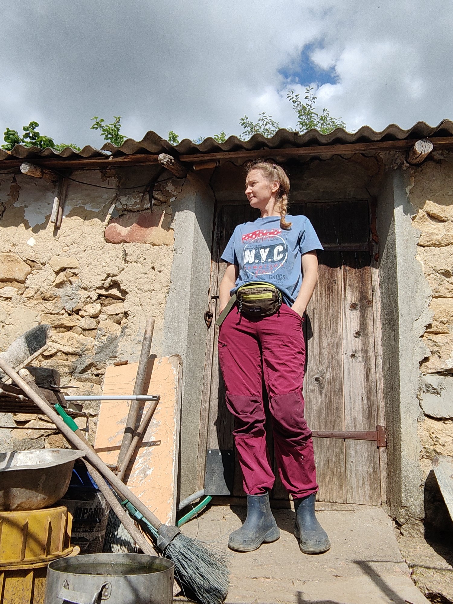 A woman stands with her hands in her pockets in front of a rustic wooden door. She is wearing a blue t-shirt with a graphic print, a green and brown fanny pack, burgundy pants, and dark rubber boots. The wall behind her is constructed of rough stones and mortar, with a corrugated metal roof overhead. To her left, several gardening tools, including brooms and a rake, are propped against the wall. The scene appears to be outdoors, with a cloudy sky visible above. The location is identified as Ароматновское сельское поселение, Ukraine.
