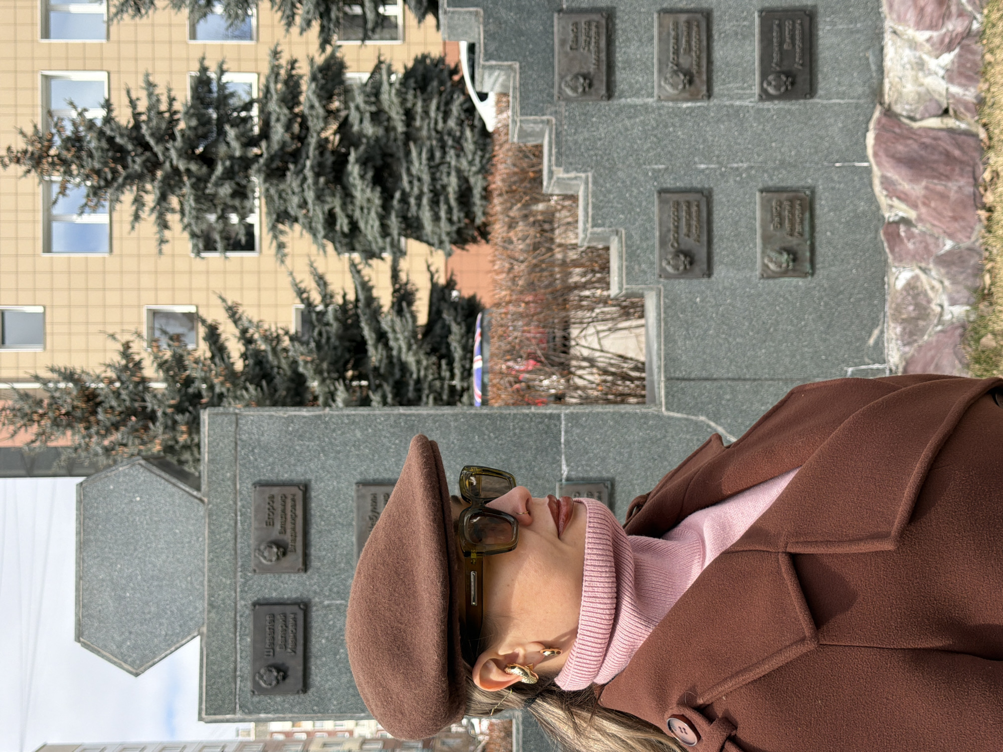 The image captures a person in the foreground against a backdrop of a memorial site, architectural structures, and trees under bright daylight. The scene suggests a public space in Krasnoyarsk, Russia.
**Subjects:** The main subject is a person, likely a woman, seen in left profile. She wears a brown beret, large dark-lensed sunglasses, a light pink turtleneck sweater, and a brown wool coat with visible buttons. She has gold-colored ear cuffs or earrings. Her gaze is directed towards the right side of the frame.
**Scene and Setting:** In the mid-ground, to the right and behind the person, stands a substantial grey granite memorial wall or stele. It features multiple rectangular bronze plaques arranged in vertical rows, each bearing a name in Cyrillic script and an embossed medallion or portrait. The memorial has tiered sections. Visible names on some plaques include "Трунова" (Trunova), "Голиков" (Golikov), "Оськин" (Oskin), and "Егор Вадимирович" (Egor Vadimirovich). Further behind the memorial is a multi-story building with a light beige or yellow grid-patterned facade and numerous rectangular windows reflecting the sky. Dark green evergreen trees, possibly spruce or fir, are prominent between the building and the memorial, while some bare deciduous branches suggest the season. A low wall constructed from reddish-brown, irregularly shaped stones borders the ground to the far right of the memorial.
**Activity or Event:** The person appears to be standing or observing the memorial, or simply present in the outdoor environment. There is no explicit activity depicted.
**Notable Details:** The image is well-lit by clear daylight. The person's attire (beret, coat, turtleneck) and the partially bare trees indicate cooler weather, consistent with autumn, spring, or a mild winter day in Krasnoyarsk. The memorial, with its numerous plaques, suggests a monument commemorating individuals, possibly heroes or significant figures.
**Visible Text:** The most prominent text is in Cyrillic script on the bronze plaques of the memorial. Legible portions include names like "Трунова", "Голиков", "Оськин", and "Егор Вадимирович".