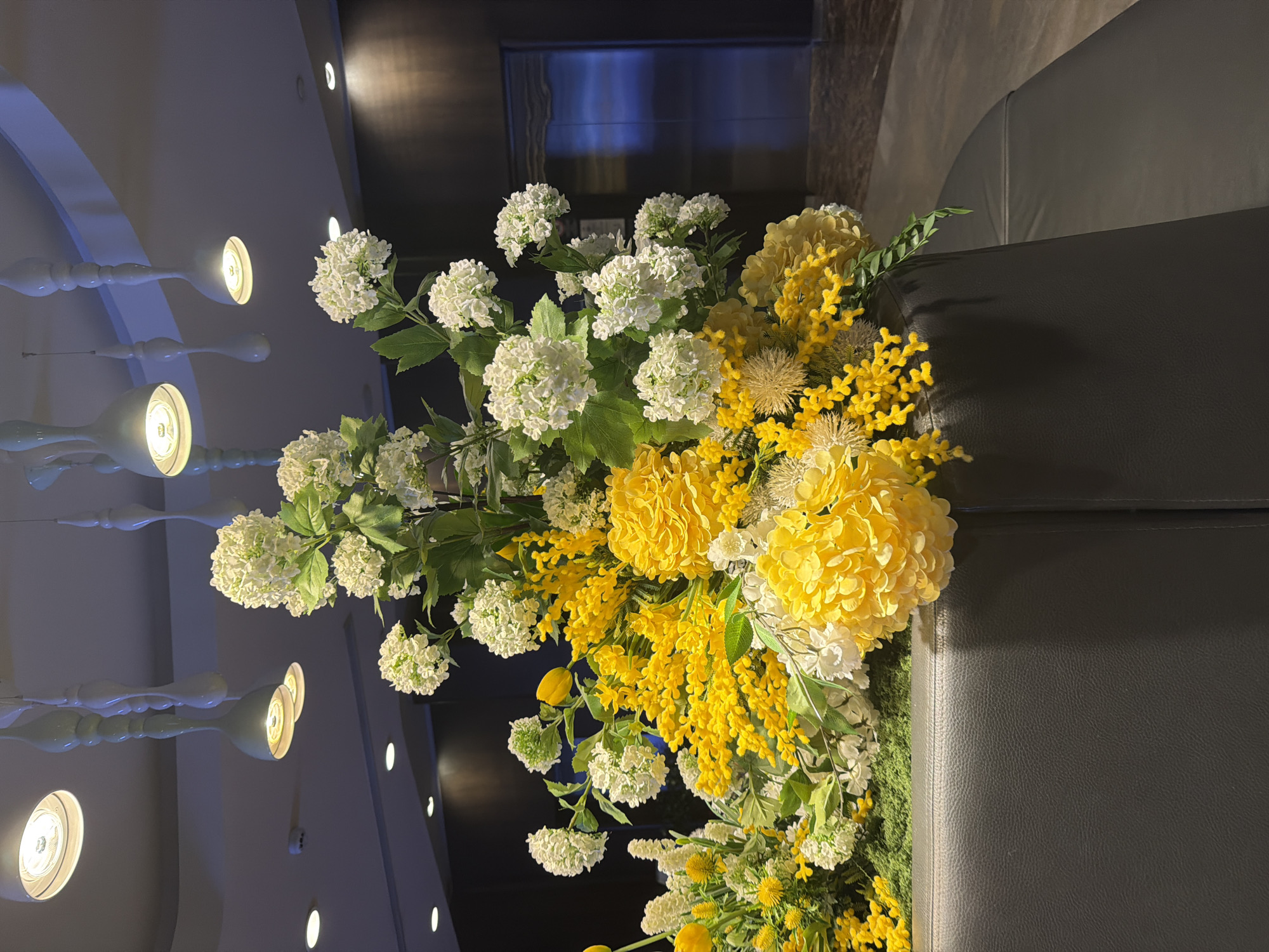 The photograph depicts a floral arrangement, combining white hydrangeas and yellow mimosa, placed atop a dark-colored piece of furniture, possibly a chair or bench. The backdrop includes elements of an interior space, such as a wall with a modern lighting fixture and part of a doorway or entryway. The scene's location, Moscow, Russia, is not discernible from the visual information provided.