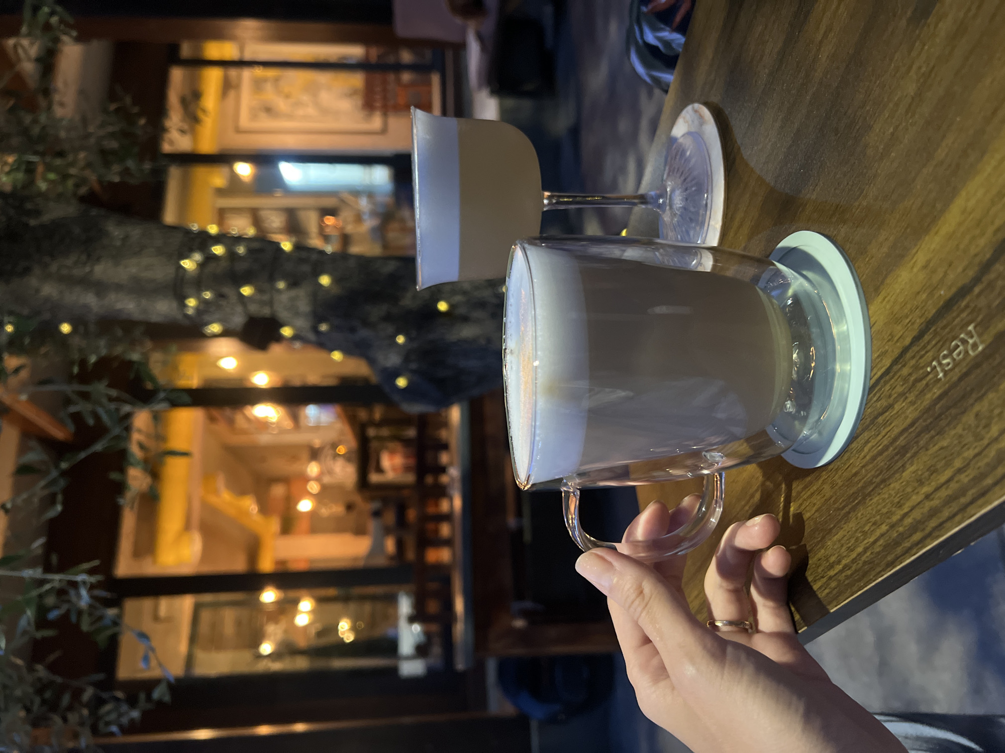 A hand with a gold ring is holding a clear mug of latte, with a foam top, on a wooden table. Next to the mug is a stemmed glass with a light-colored drink and a white foam top. In the background, a tree is decorated with fairy lights, and the blurred lights of a building are visible. The scene appears to be outdoors, possibly a cafe or restaurant, in Bjeliši, Montenegro.