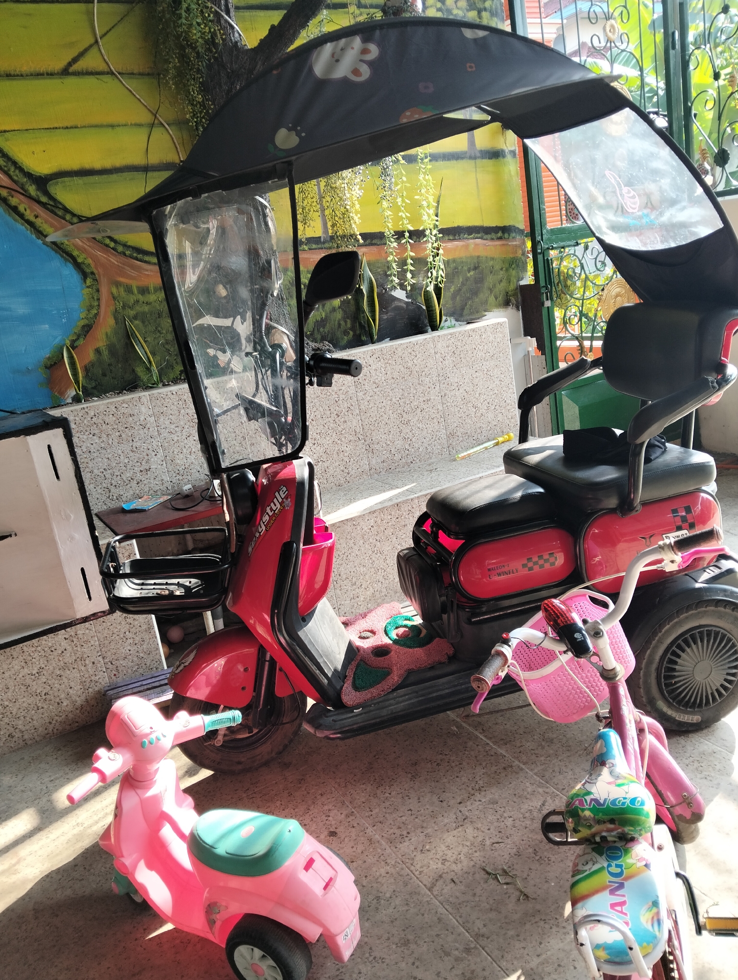 In Muncar, Indonesia, a bright pink toy scooter sits on a tiled floor in front of a larger red electric scooter. The toy scooter has a green seat and handlebars. The electric scooter is equipped with a black canopy and a clear plastic windshield, and has a black seat. A smaller pink bicycle with a basket is parked beside the electric scooter. Behind the scooters, a wall features a colorful mural depicting a body of water and a landscape. The scene is illuminated by sunlight, casting shadows on the floor.