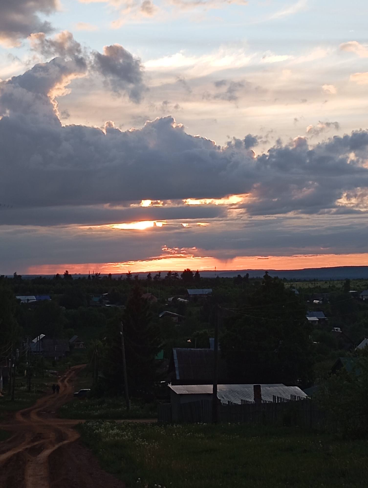 The image depicts a sunset view over a rural landscape. The sky is filled with layered clouds, ranging from dark gray to light blue, with streaks of orange and yellow where the sun is setting. The horizon is a line of trees and vegetation. In the foreground, there is a dirt road winding through a small settlement with houses and fences. Two individuals are walking on the road. The location is Фоки, Russia, as specified in the query.