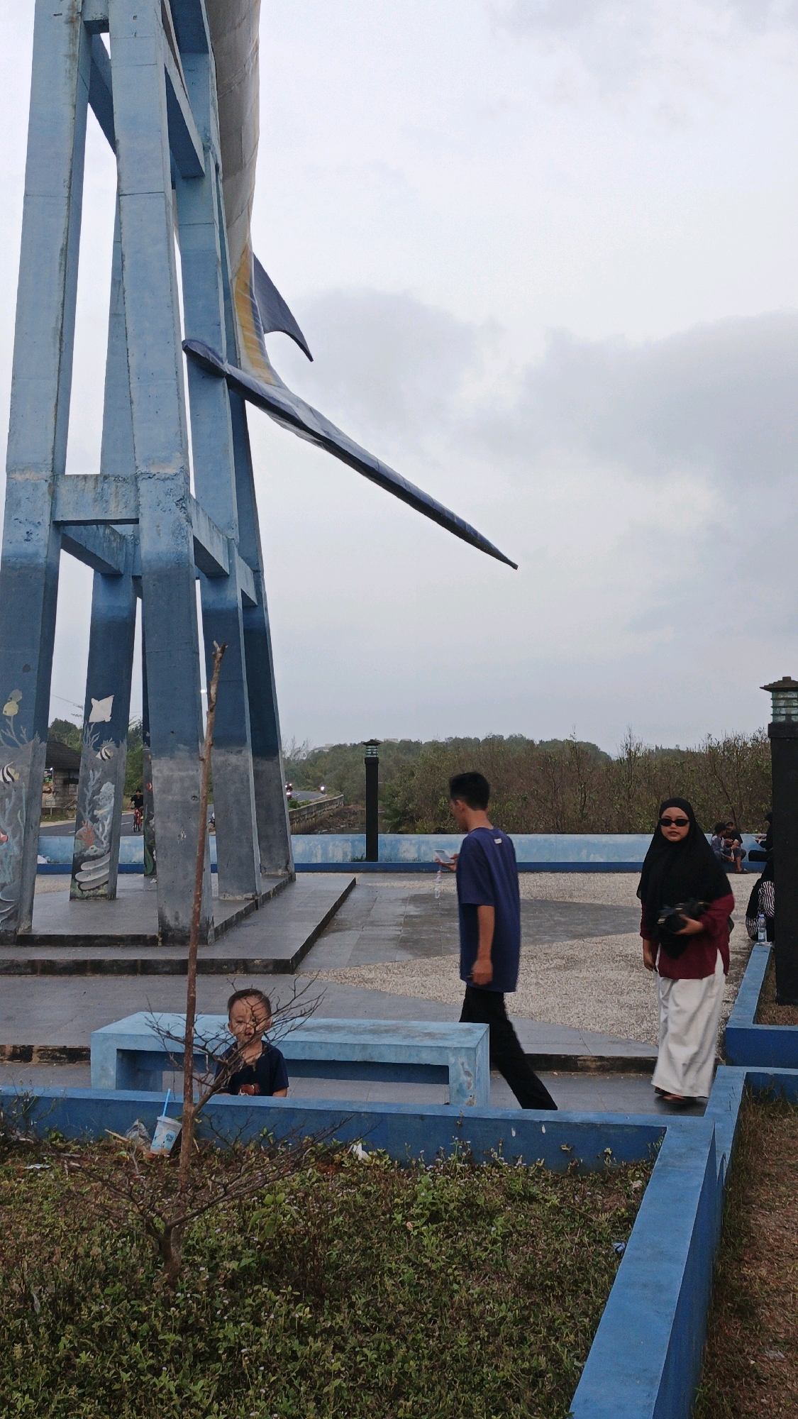 The image depicts a monument structure in Pangandaran, Indonesia, characterized by large, blue, concrete pillars supporting a stylized, dark-colored fish sculpture, possibly a marlin. The fish is positioned horizontally, with its tail and fin elements visible. The surrounding area features a paved plaza with blue benches and low-lying greenery.
In the foreground, a young child is seated on a blue bench, partially obscured by foliage and a thin tree branch. The child is wearing a dark t-shirt and appears to be looking towards the camera.
Further into the scene, two individuals are walking across the plaza. A young man in a dark blue t-shirt and black pants walks from right to left, holding a phone. To his right, a woman wearing a black hijab and sunglasses is dressed in a maroon top and a white skirt. She is also walking from right to left, holding a black bag.
In the background, a road is visible with some passing vehicles. The landscape beyond the plaza is characterized by dense, dry vegetation and trees under an overcast sky. The overall atmosphere suggests a public outdoor space, possibly a park or tourist attraction. The monument appears to be a significant landmark in the area.