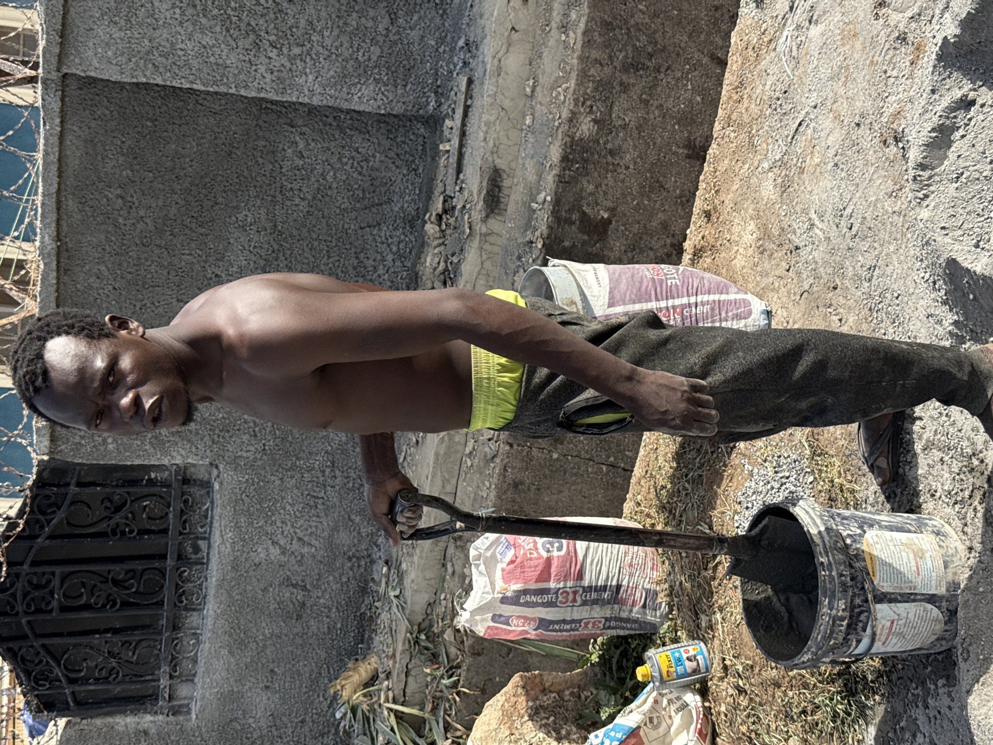 The image, captured in Abuja, Nigeria, shows a shirtless man engaged in manual labor at what appears to be a construction or renovation site during bright daylight. The man, with dark, short curly hair, looks directly at the camera with a neutral to serious expression. He wears dark trousers over bright yellow-green shorts.
He is holding a shovel with both hands, its blade submerged in a black bucket filled with a dark, mixed substance, likely concrete or mortar. This suggests he is in the process of mixing or moving building materials.
Around him on the rough ground are two bags of cement: one purple bag clearly labeled "DANGOTE CEMENT (42.5)" and another white and red bag bearing the text "DANGOTE 3X CEMENT" and "NG CD". A small yellow-capped plastic bottle, labeled "FIXIT" and "101 W+", sits near the cement bags, possibly an additive for the mix.
In the background, solid concrete walls frame the scene. To the left, an ornate dark metal gate or window grill is visible, partially obscured by barbed wire, indicating a secure boundary. The strong shadows and clear visibility suggest it is midday or early afternoon on a sunny day.
