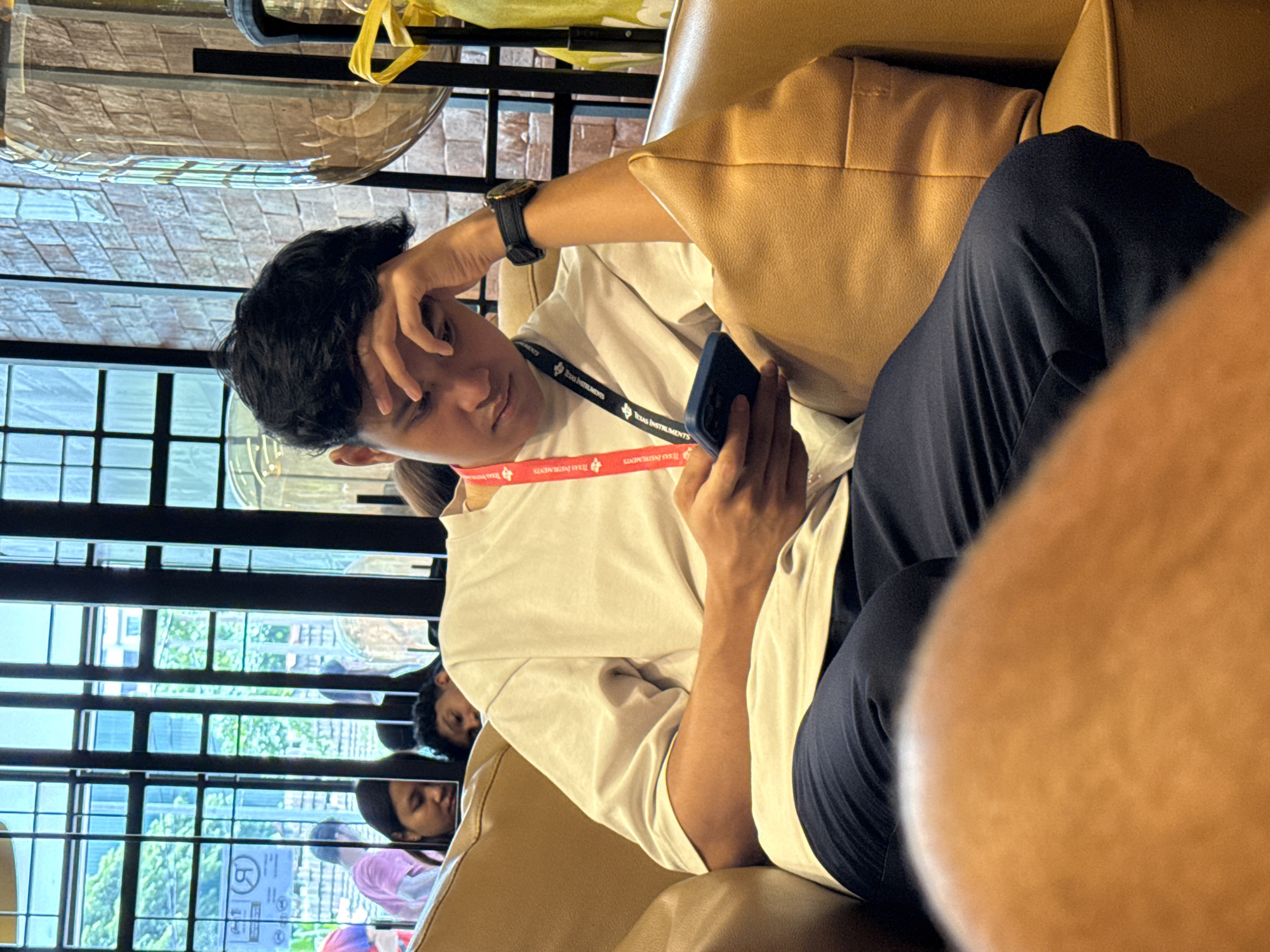 A young man is seated in an armchair with tan leather upholstery. He is wearing a white t-shirt, navy blue pants, and a watch on his left wrist. He is holding a mobile phone in his right hand and touching his forehead with his left hand, appearing either stressed or deep in thought. A lanyard with "TEXAS INSTRUMENTS" printed on it hangs around his neck. The background shows a window with multiple horizontal panes, outside of which there are trees and buildings. Additional people are partially visible behind him. The location is potentially Kuala Lumpur, Malaysia, based on the window view and the style of the chair.