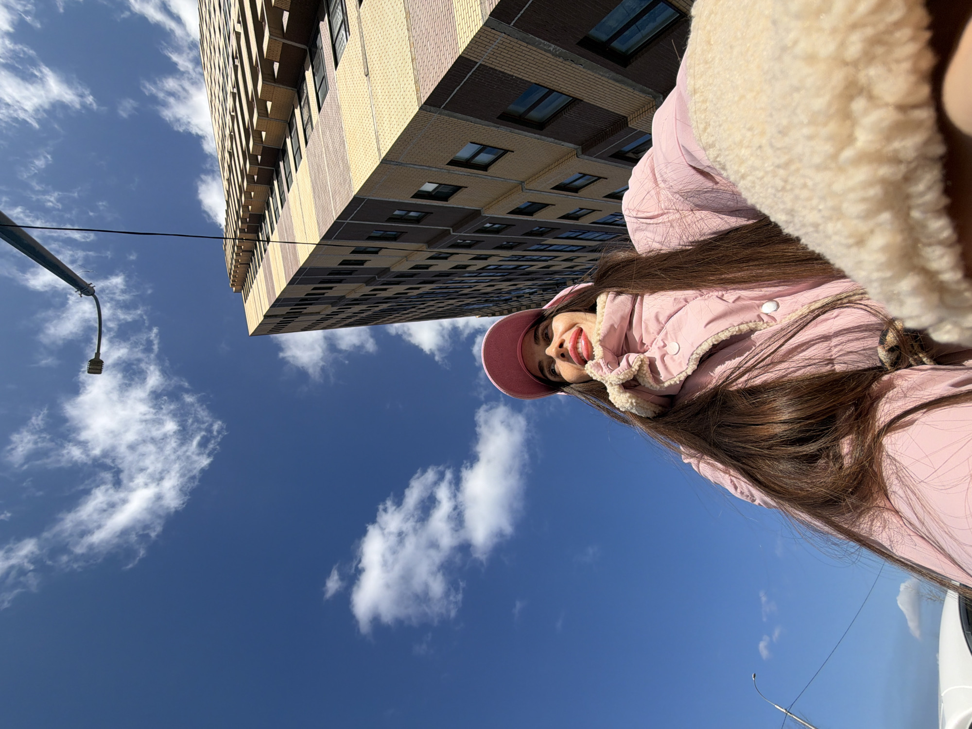 A low-angle outdoor selfie captures a single female subject against the backdrop of a tall, modern residential building in Saint Petersburg, Russia, specifically identified as LEGENDA na Geroyev 24/2, корпус 1, in the Krasnoselsky District. The scene is illuminated by bright sunlight under a clear blue sky dotted with scattered white cumulus clouds.
The female subject, positioned in the lower-center of the frame, has long, dark brown hair and is wearing a pink baseball cap, a light pink puffer jacket with visible cream-colored sherpa lining on the collar/hood and right cuff, and red lipstick. She gazes upwards and to her left with a slight smile. Her right arm, implied to be holding the camera, is partially visible and covered by the jacket's sherpa cuff on the right edge of the frame.
The background is dominated by a multi-story building rising sharply upwards on the right side, exhibiting a modern architectural style with alternating horizontal bands of light beige/yellow and dark brown brick-like facade panels. Numerous dark-framed rectangular windows are visible across its surface. In the upper-left, a black street light pole extends diagonally across the sky, equipped with a luminaire and a hanging black cable. A partial view of the white roof of a vehicle is present in the lower-left corner.