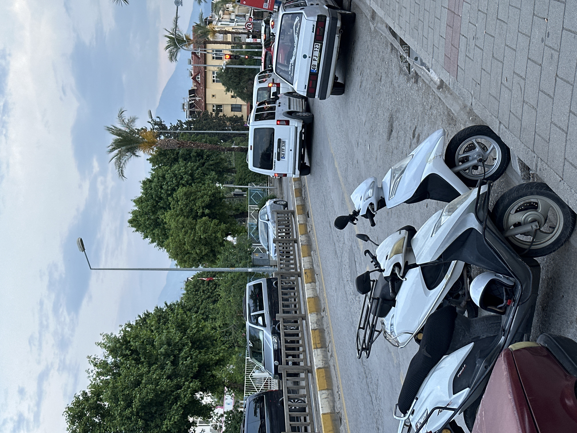 The image shows a street scene in Fethiye, Turkey. Several vehicles are parked or stopped on the road, including white vans, a white car with license plate "48 PJ 123", and other cars. Several scooters, including white ones, are parked on the sidewalk. A white helmet is visible on one of the scooters. The license plate of a van reads "48 FF 538". Palm trees and other greenery line the street. Buildings are visible in the background, and mountains can be seen in the distance under a cloudy sky. A street light is also visible. The scene appears to be a typical street in a residential or commercial area of Fethiye.