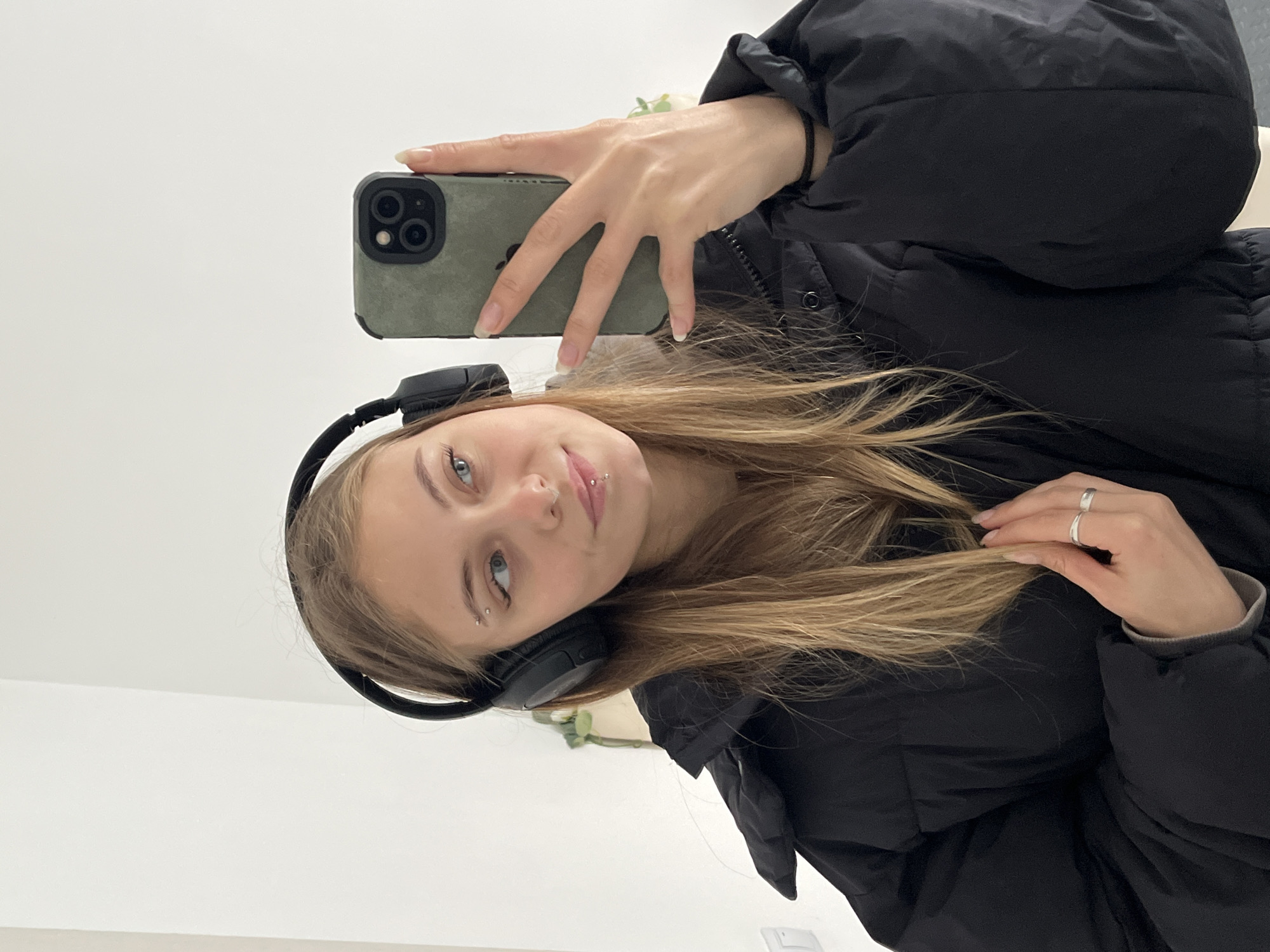 A young woman takes a selfie in what appears to be an indoor location. She wears a black puffer jacket and black over-ear headphones. She holds a mobile phone with a green case in her left hand, the screen facing away from the camera. Her long, blonde hair is partially covering her face. She has multiple piercings in her face: one in her eyebrow, one in her nose, and one in her lip. She also wears two rings on her right hand. The background is a plain, light-colored wall. There is no evidence of the location being in Фоки, Russia.