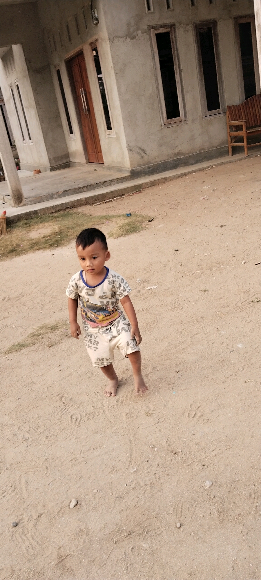 A young boy, appearing to be of Southeast Asian descent, stands on a sandy ground in front of a concrete house. He has dark hair and is wearing a light-colored t-shirt with a graphic print of a car and letters, and matching shorts. His left foot is slightly forward, and his right leg is bent, suggesting he is in motion. The house in the background has a light-colored concrete facade, a wooden double door, and several tall, narrow windows with wooden frames. To the right of the house, a wooden bench is partially visible. The ground is a mix of sand and some scattered debris, with a hint of dry grass to the left of the boy. The lighting suggests it is daytime. This scene is likely located in Tanjung Ratu Ilir, Indonesia, a region characterized by its coastal geography and tropical climate, which would explain the sandy terrain and typical residential architecture.
