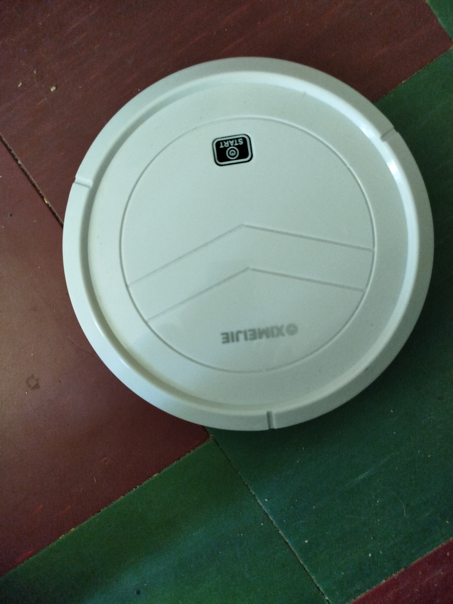 A white, circular robot vacuum cleaner rests on a floor with alternating dark red and green sections. The robot has a black rectangular button with a white "START" label and a circular icon inside. The brand name "XIMELJIE" is printed in white text on the robot's surface. The scene appears to be indoors, and no people are visible. The lighting suggests it is daytime. The location in Pavlohrad, Ukraine, is not visually ascertainable from the image alone.