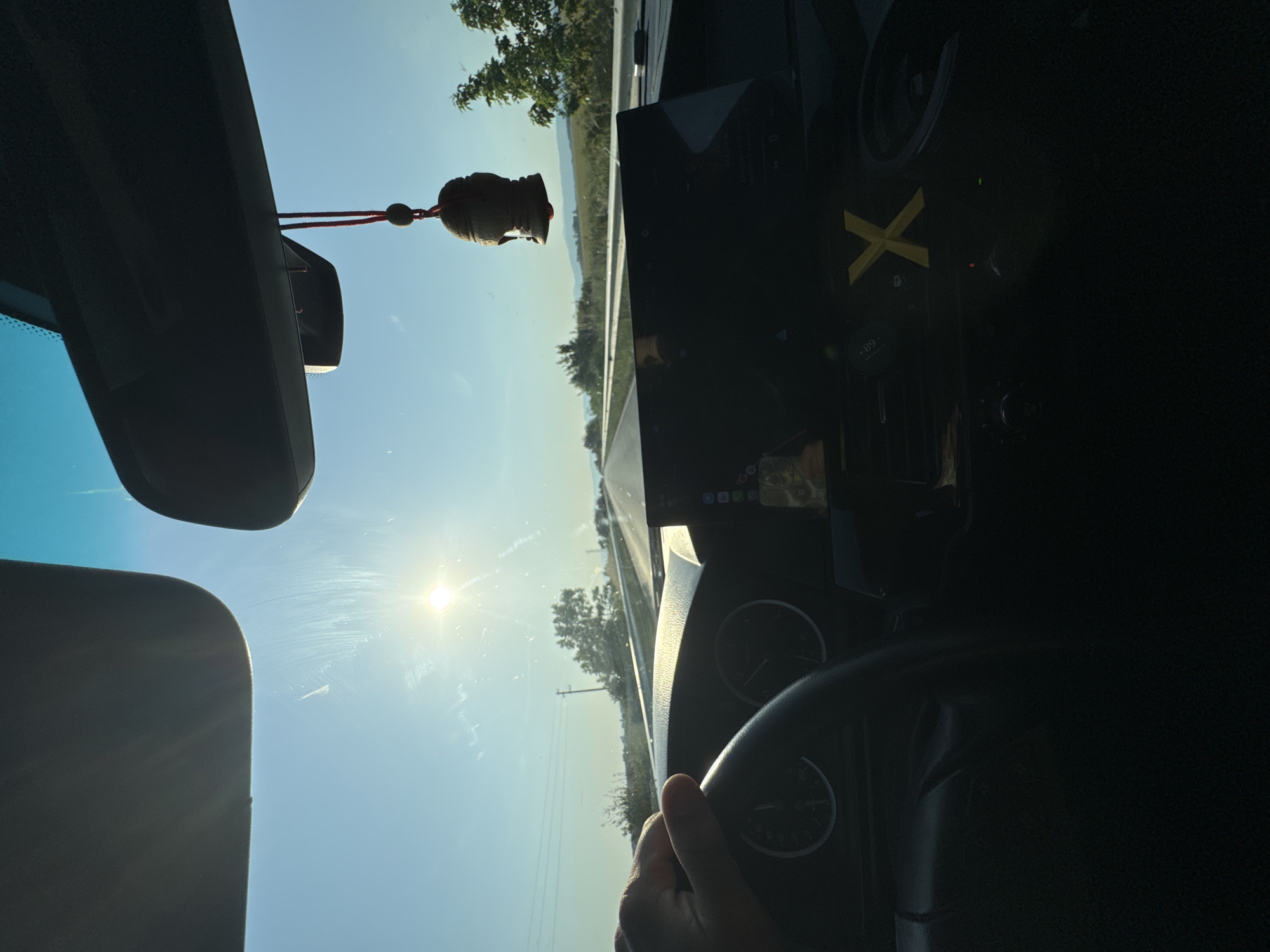 The image presents a first-person perspective from the interior of a vehicle, traveling on a road in the vicinity of Mogila, Bulgaria, under a clear sky. The sun is low in the upper-left quadrant of the frame, shining directly through the windshield and causing significant lens flare.
Within the vehicle's interior, a gray sun visor is partially visible in the upper-left corner. A dark rearview mirror assembly, encompassing sensor housing, is centrally positioned at the top of the frame. A small, brown, carved ornament, resembling a Buddha head, hangs from the rearview mirror by a red cord.
A light-skinned left hand is visible, gripping the black steering wheel on the left side of the frame. Behind the steering wheel, parts of the instrument cluster are discernible, showing numerical increments reaching at least 260. The dashboard features a prominent, dark central display screen. Below this screen, a distinct yellow letter "X" is affixed to the dashboard, with a small digital display showing ".69" to its left.
The exterior environment depicts an asphalt road extending into the distance. A metal guardrail lines the right side of the road, beyond which green vegetation and fields are visible. Scattered trees and bushes populate the mid-ground, and a utility pole stands on the left side of the road. Distant, hazy hills or mountains form the horizon line under a blue sky, which lightens towards the sun.