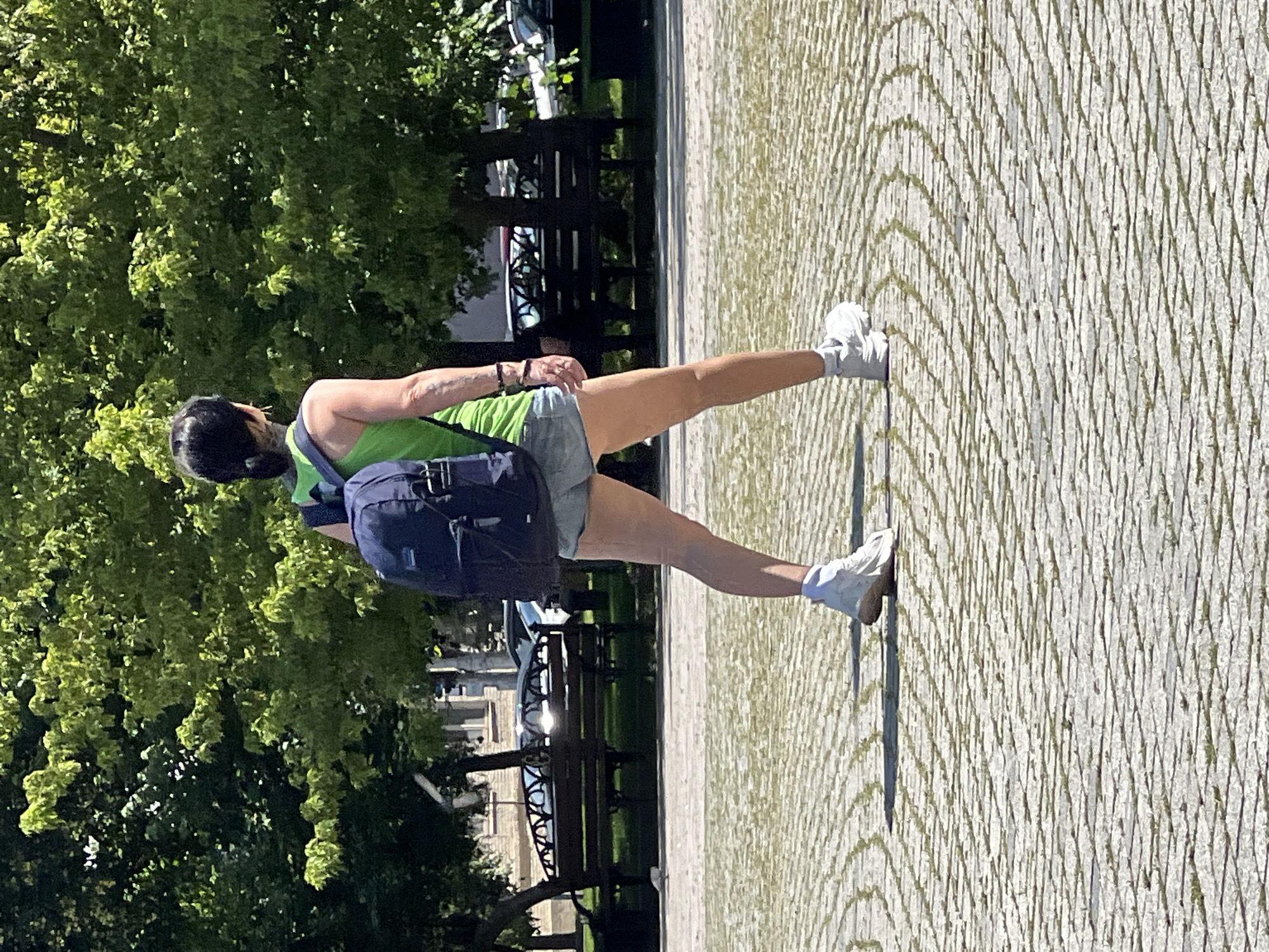 A person wearing a green tank top, denim shorts, and white sneakers walks across a paved plaza. The person has a dark ponytail and is carrying a dark blue backpack. In the background, there are trees, park benches, and some cars. The scene appears to be a sunny day in a public park or square. The specific location in Krasnodar, Russia cannot be determined from the provided image.