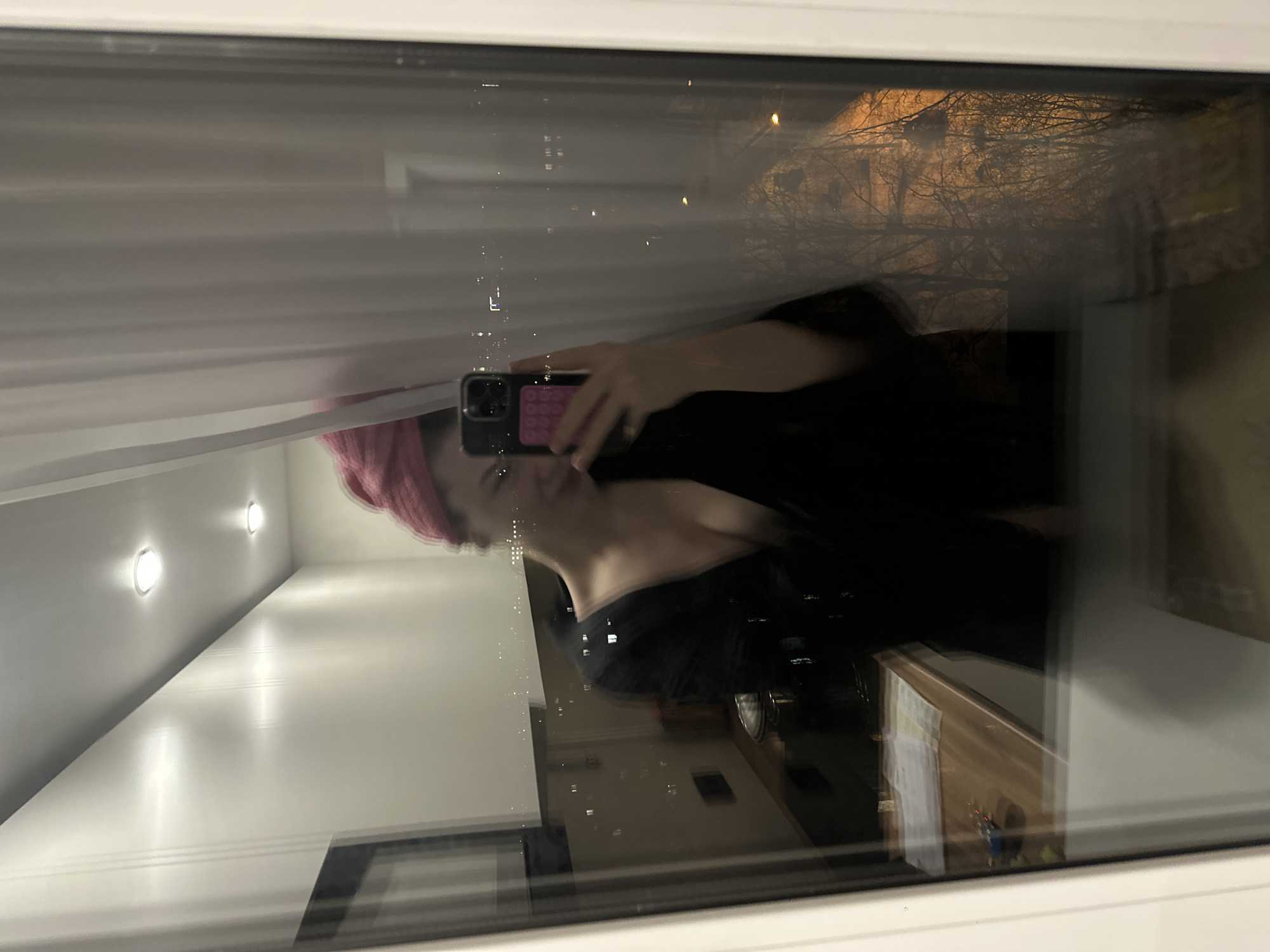 A woman with pink hair and a black top is taking a selfie in a window at night. The reflection in the window shows the interior of a room with a light-colored ceiling and recessed lighting, as well as an exterior view of what appears to be buildings and trees against a dark sky. Based on visual clues of Cyrillic signs, the location is estimated to be in Kazan, Russia.