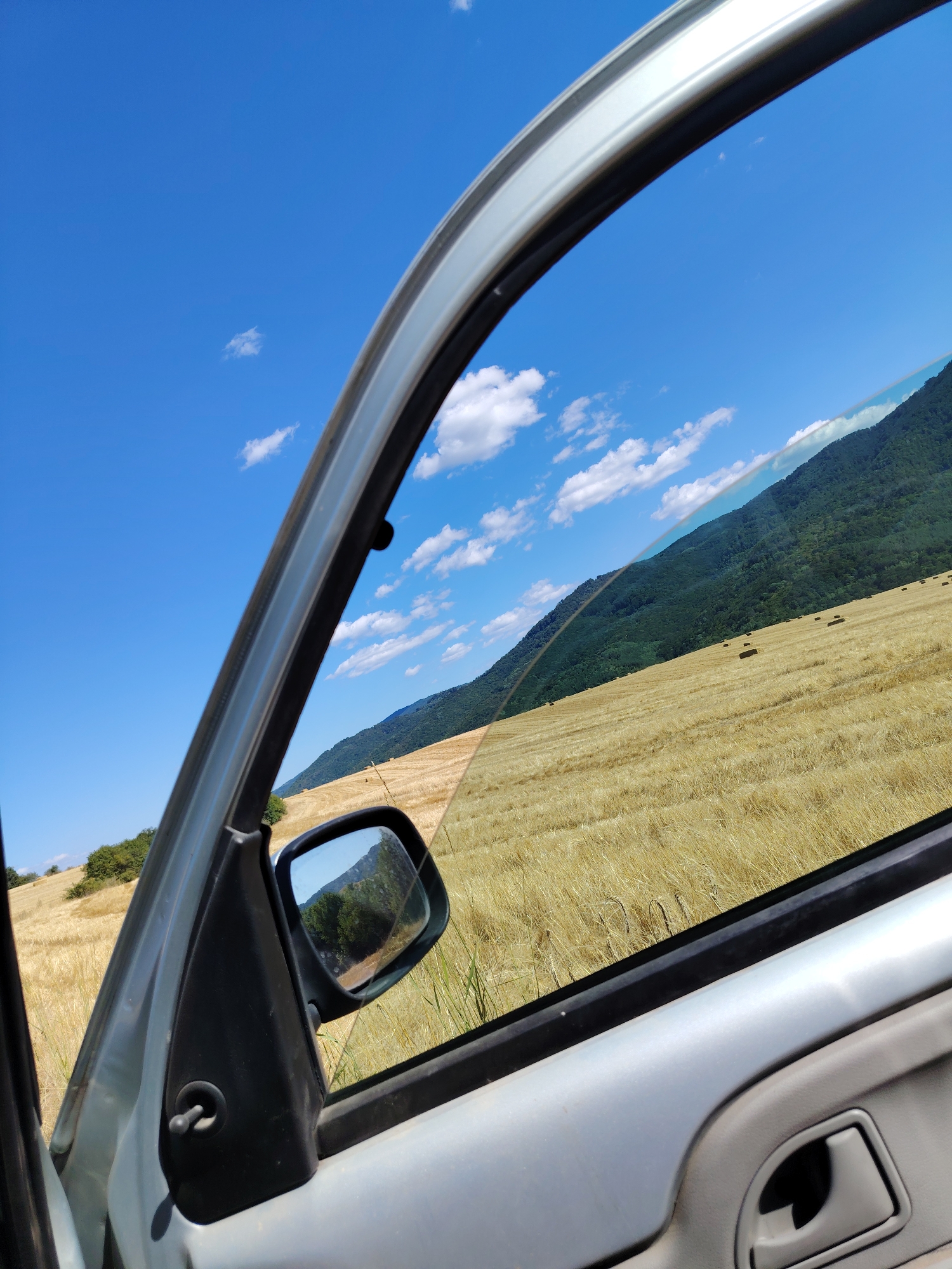 The image, taken from inside a vehicle, showcases a rural landscape. The foreground is dominated by the interior of a car door, with the window framing the exterior view. A side-view mirror is visible, reflecting a section of the landscape. The exterior scene features a vast field of golden, dry grass, possibly harvested wheat or barley, with scattered square bales of hay. In the background, a lush green, tree-covered mountain slopes upwards against a clear blue sky dotted with white, fluffy clouds. The location is identified as Dushantsi, Bulgaria.